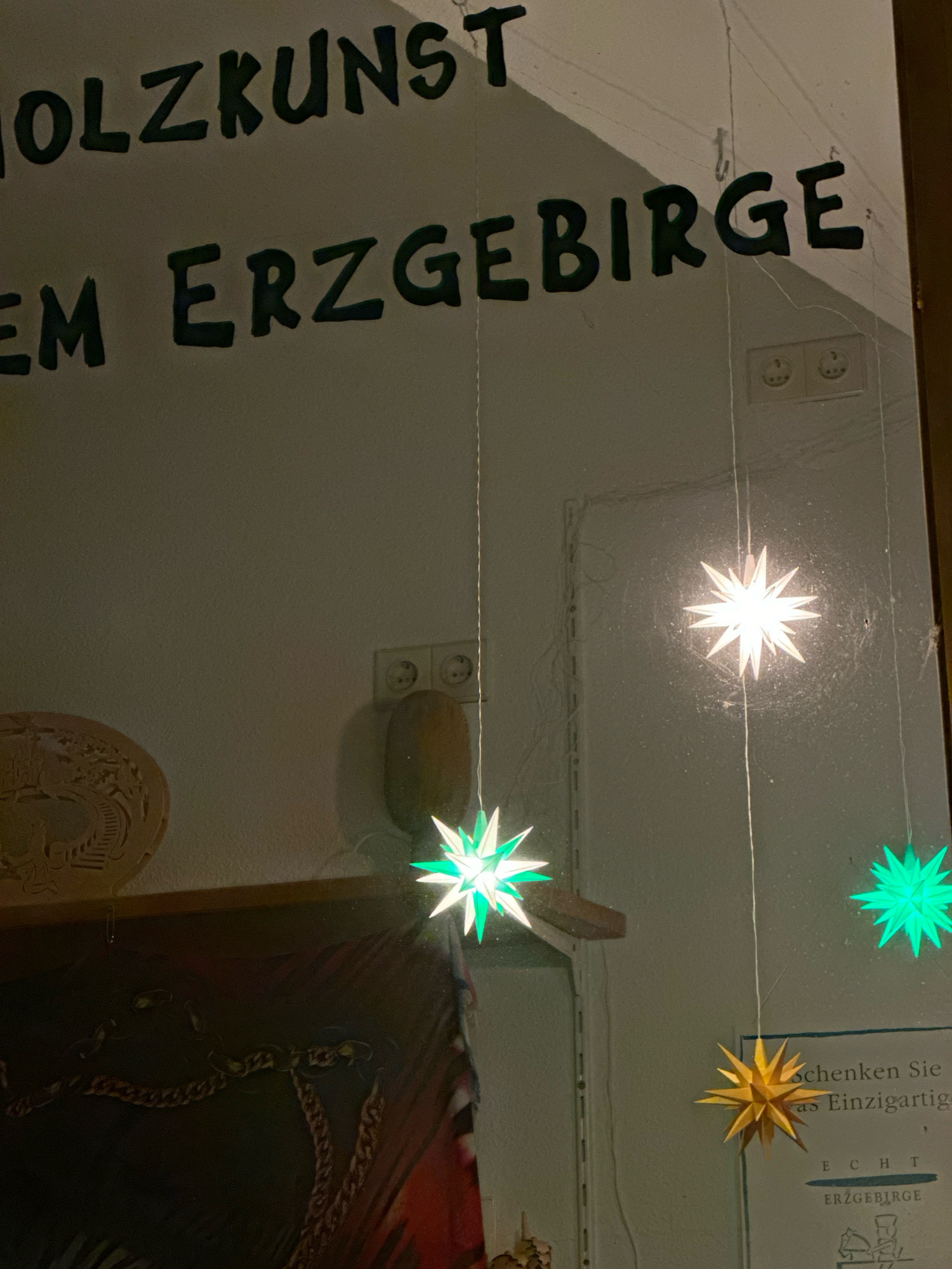 The image shows a shop window display. There is German text written on the window, which appears to read "HOLZKUNST" at the top and "EM ERZGEBIRGE" below it. Several paper stars are hanging in the window, some are lit up and glowing. One star is white, another is green, and a third is yellow. A sign with text in German is visible in the lower right corner, and the word "ERZGEBIRGE" is clearly legible. The shop appears to sell wooden crafts from the Erzgebirge region of Germany, known for its traditional folk art and Christmas decorations. The overall impression is of a festive and inviting display. The specific location in Krefeld, Germany is not provided in the image.