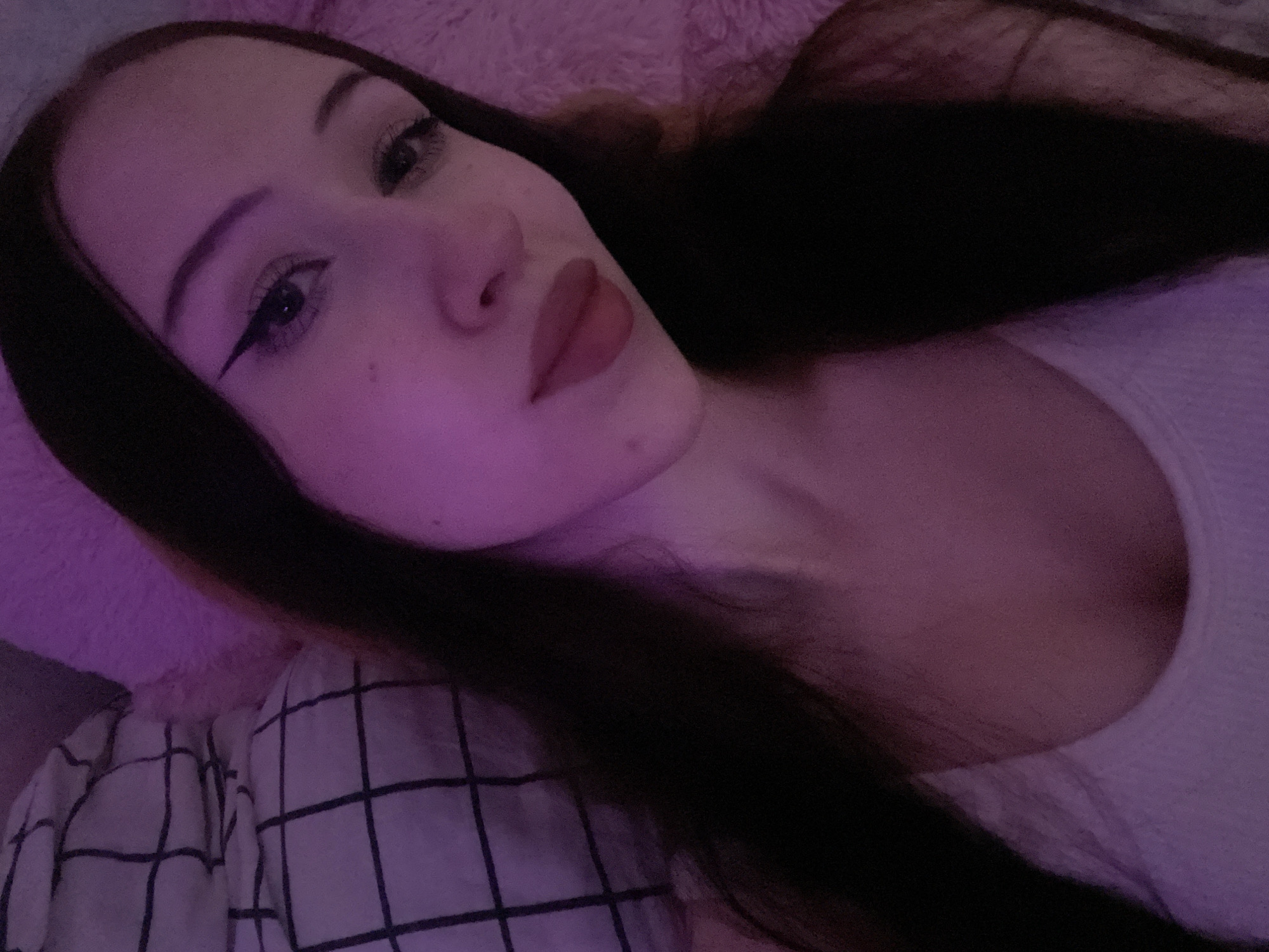 A medium shot shows a young woman with long, dark hair and fair skin. She is lying down, with her head tilted slightly. The lighting is dim and tinted pink, creating a soft glow. She is wearing a white tank top. Her eyes are lined with black eyeliner, and she has pink lipstick on. Behind her, a pillow with a black and white checkered pattern is visible, along with a fluffy pink surface, possibly a blanket or another pillow. There is no indication of the city of Votkinsk, Russia in the image.