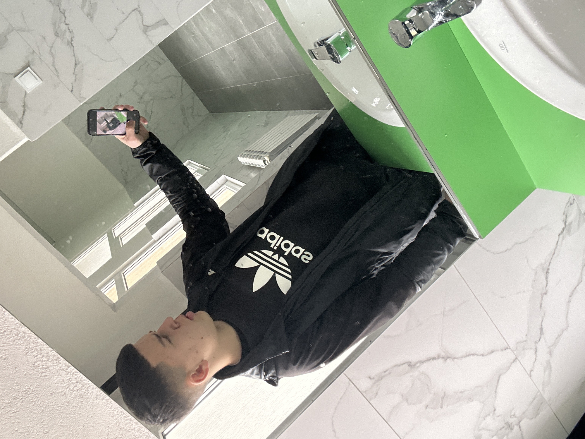 A young man with short dark hair is taking a selfie in a bathroom. He is wearing a black Adidas T-shirt and a black jacket. The bathroom has marble walls and floors, a green sink, and a window. The location may be in Zlatograd, Bulgaria.