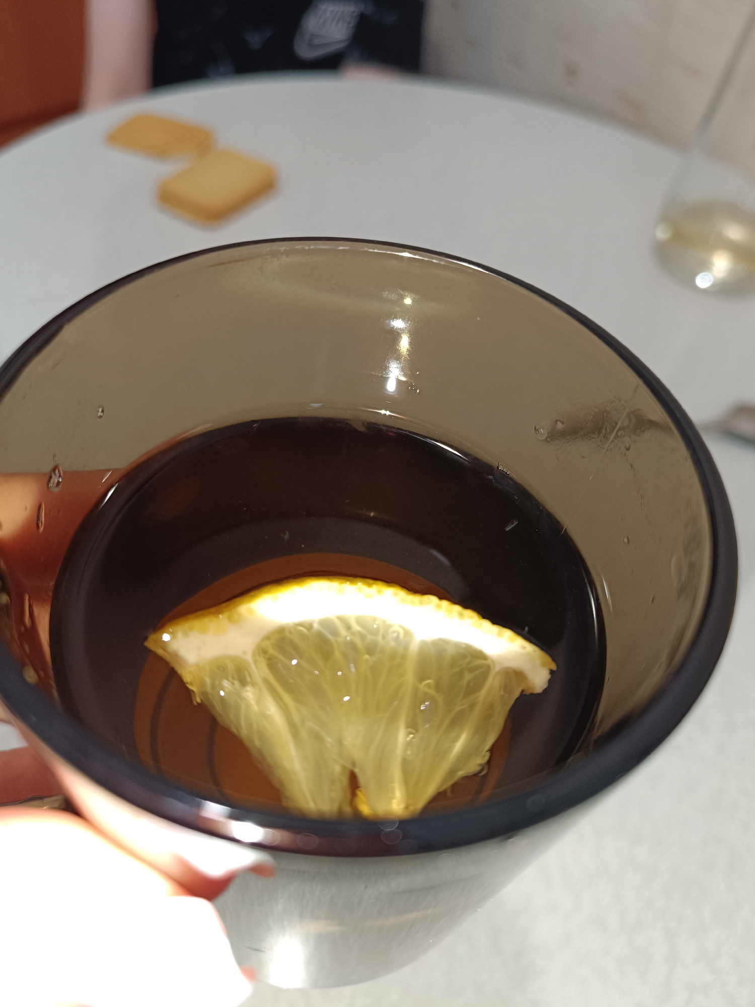 A close-up shot depicts a dark-colored beverage in a tall, tinted glass. A slice of lemon floats inside the drink. In the blurred background, two square biscuits are on a white table. Part of another glass with a clear liquid is also visible. The image contains a person's hand holding the glass, part of another person seated at the table. The scene does not provide information that would identify it as being specifically located in Tobolsk, Russia.