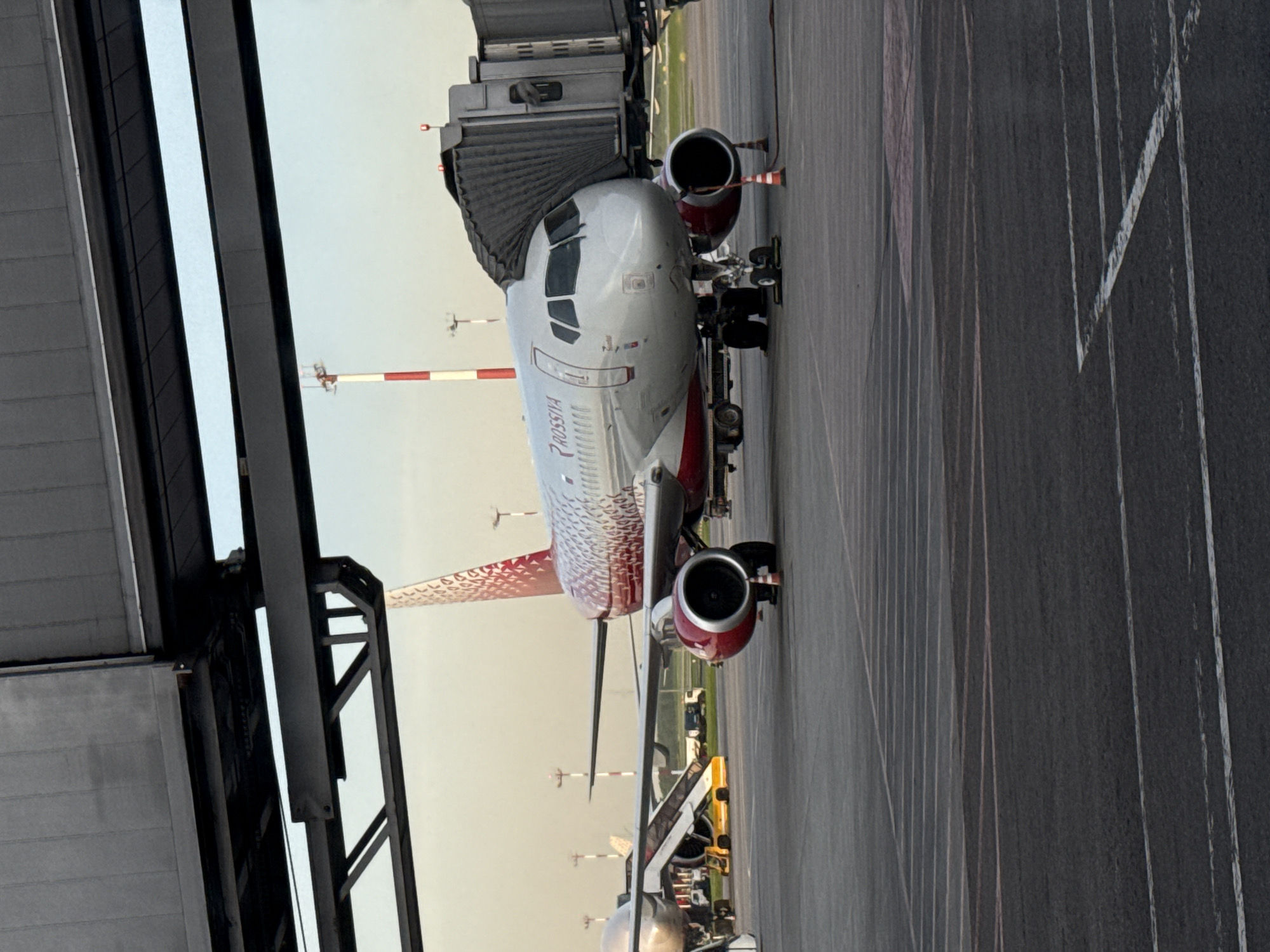 The image depicts a Rossiya Airlines aircraft parked at a terminal in Khimki, Russia. The plane is connected to a jet bridge, and ground service vehicles are nearby. The environment appears to be an airport tarmac, with visible runway markings.