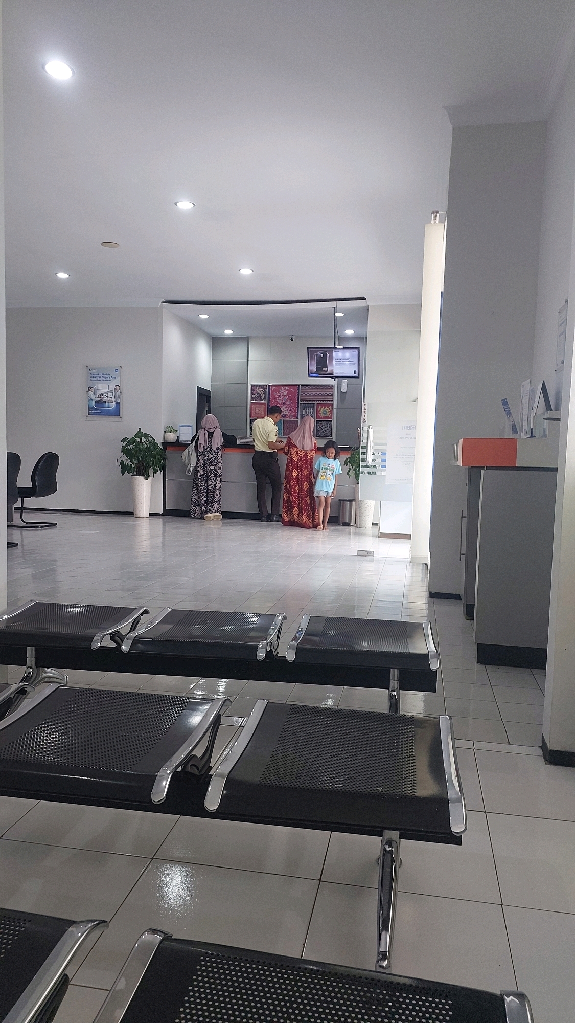 The image depicts the interior of a government office, likely a post office or similar administrative building, in Sukowono, Indonesia. The scene is characterized by a clean, tiled floor and a well-lit environment with overhead fluorescent lighting. In the foreground, rows of empty black metal chairs with chrome accents suggest a waiting area.
Further back, a reception counter is visible with several individuals. Two women in traditional Islamic attire are standing near the counter. One woman, on the left, is wearing a patterned abaya and a hijab. The other woman, to her right, is wearing a vibrant red and orange patterned dress and a hijab. Between them, a man in a light yellow collared shirt and dark trousers appears to be interacting with them, possibly an employee. To the right of this group, a young girl in a light blue t-shirt and shorts stands near the counter, looking towards the right.
Behind the counter, a television screen displays some form of information, and various posters and decorative items are visible on the wall, including some colorful woven textiles. A potted plant adds a touch of greenery to the space. The overall impression is one of an orderly and functional public service environment.