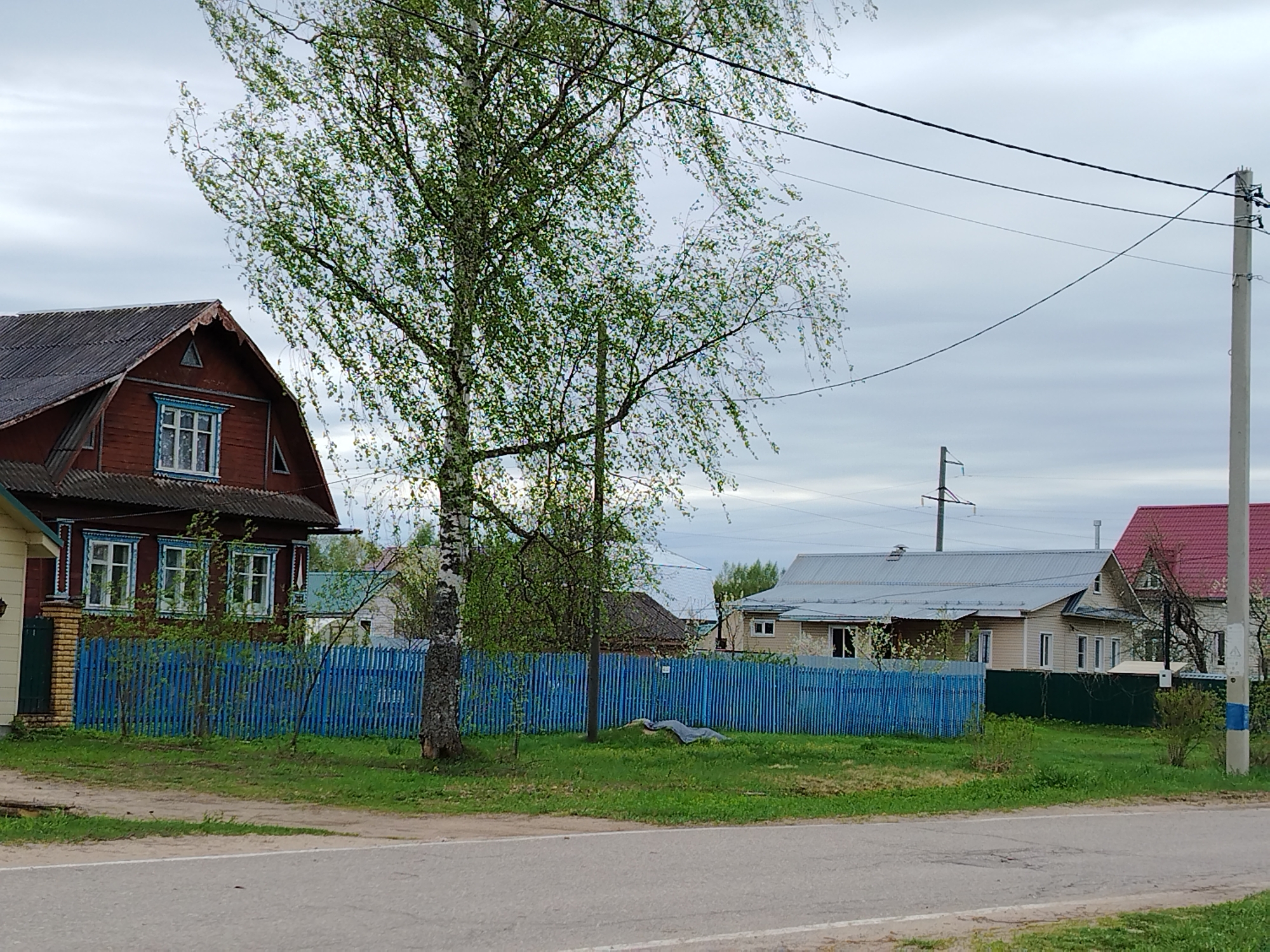 The image shows a street scene in a town, with a road in the foreground. On the left is a two-story, brown-painted wooden house with white-trimmed windows and a dark roof. A yellow-painted structure abuts the side of the brown house. A bright blue wooden picket fence runs along the property line, extending to the right. Behind the fence is a grassy area with a light-colored object lying on the ground. Trees with light green leaves are scattered throughout the scene. In the background are several other houses with various colors of roofs. Power lines are visible stretching across the sky. A light-colored concrete utility pole stands on the right side of the image. The sky is overcast.