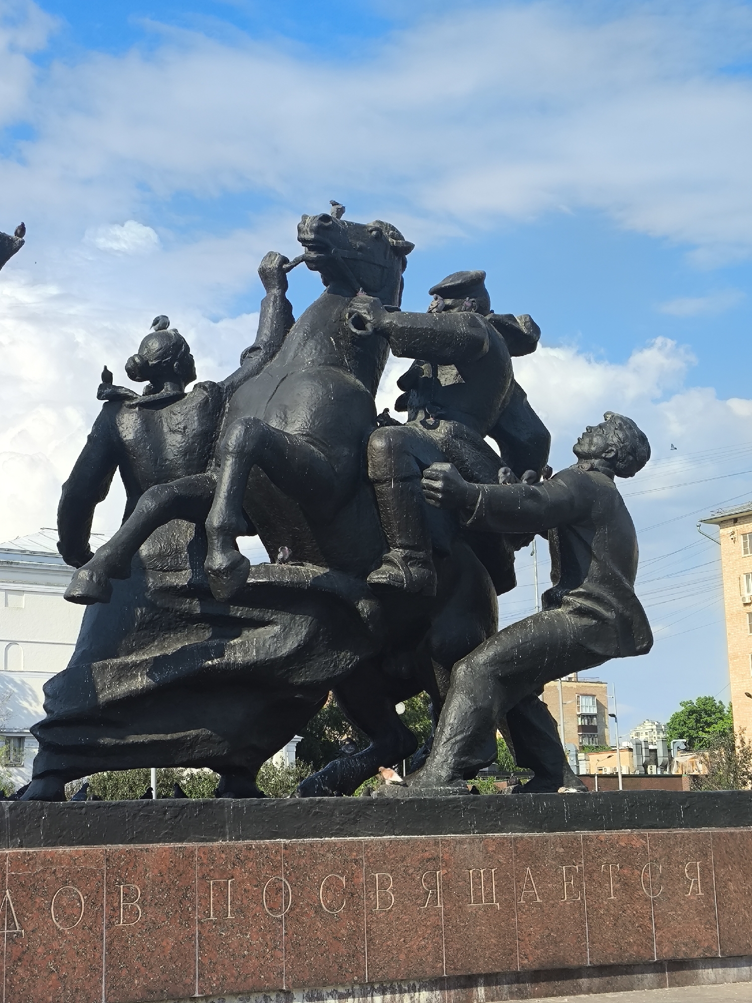 The image captures a bronze monument in an urban setting, likely in Moscow, Russia. The monument depicts a dynamic scene with multiple figures centered around a horse. Three individuals appear to be assisting another person onto the horse, while a fourth individual is already mounted. The figures are rendered in a style reminiscent of socialist realism. Pigeons perch on different parts of the statue. The monument stands on a marble base with inscriptions in Cyrillic script, parts of the words are visible including "дов посвящается" (devoted to). In the background, there are buildings with architecture typical of Russian cities, along with some greenery. The sky is blue with scattered white clouds. Electrical lines can be seen stretching across the top right of the frame. The overall impression is of a historical scene rendered in a monumental style.