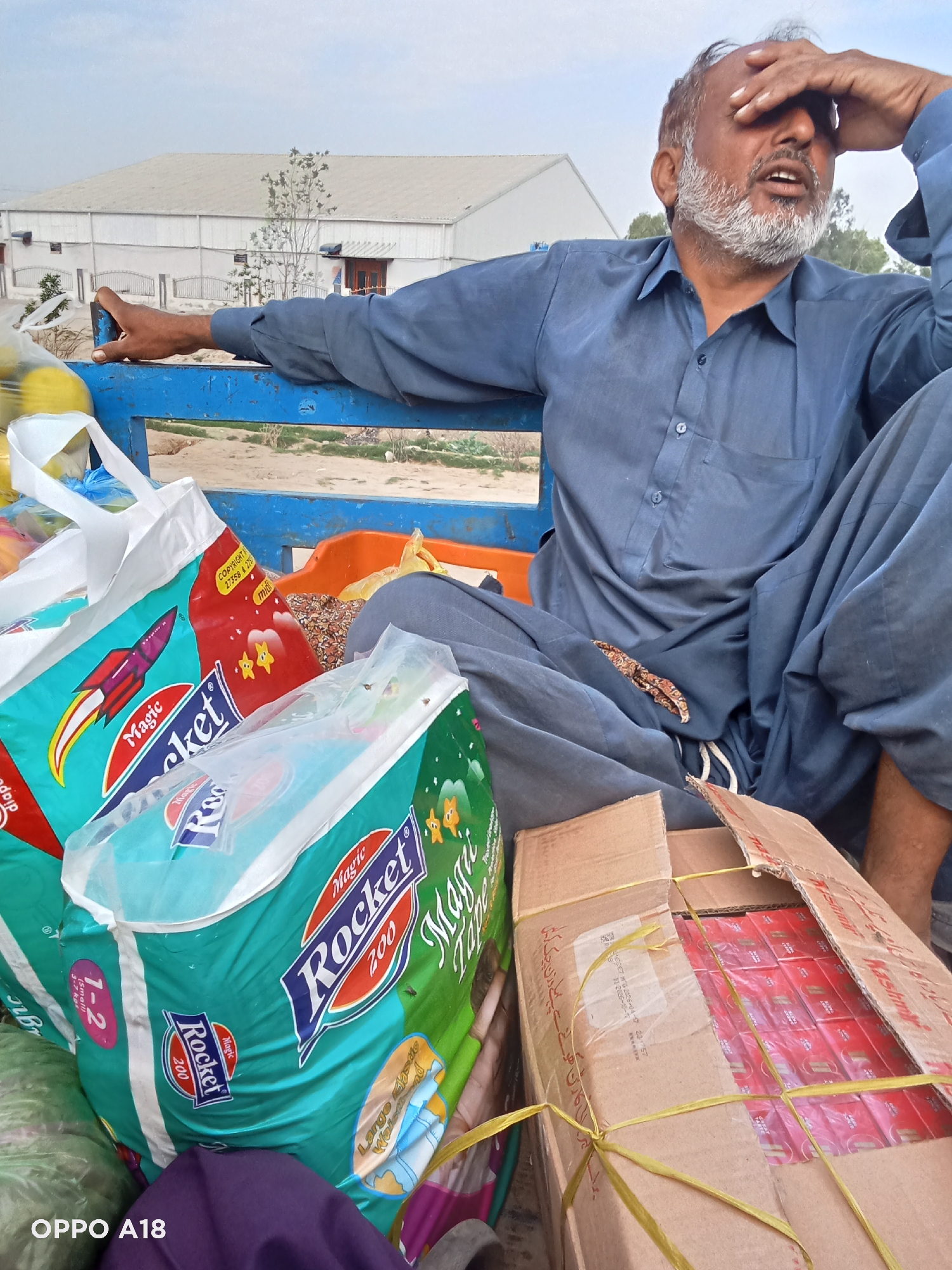 A medium shot depicts an older man of Asian descent sitting on a blue metallic structure, possibly the back of a truck or cart, filled with goods. The man is dressed in a loose-fitting blue shirt and pants, with his right hand raised to shield his eyes. He has a gray beard and appears to be looking upwards and slightly to the right.
In front of him are multiple bags of "Magic Rocket" diapers, in red and green packaging. There is also a cardboard box filled with red packets of what appears to be "Kashmir" cigarettes. These items suggest he may be involved in some form of commerce or trade. The environment suggests an outdoor setting, perhaps a rural area in Unknown, Unknown, with a large white building in the background.
