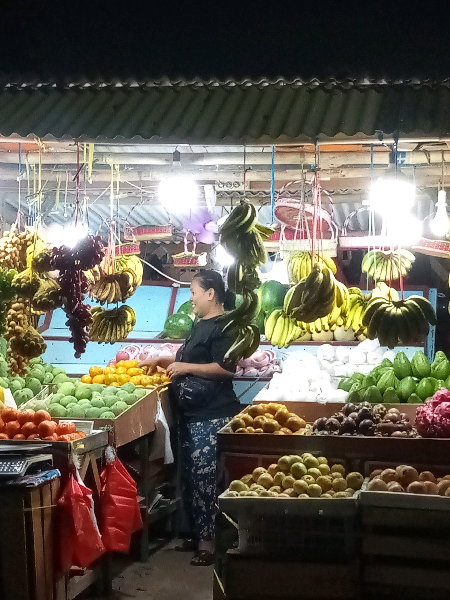 A woman is standing at a fruit stall at night in Bekasi, Indonesia. The stall is illuminated by overhead lights and displays a variety of fruits, including bananas, grapes, tomatoes, mangoes, limes, and other produce. The woman, wearing a black t-shirt and patterned pants, is interacting with the fruit, possibly arranging it or preparing to sell it. The background shows the corrugated metal roof of the stall and other parts of the market. The scene suggests a typical street market environment in Indonesia.