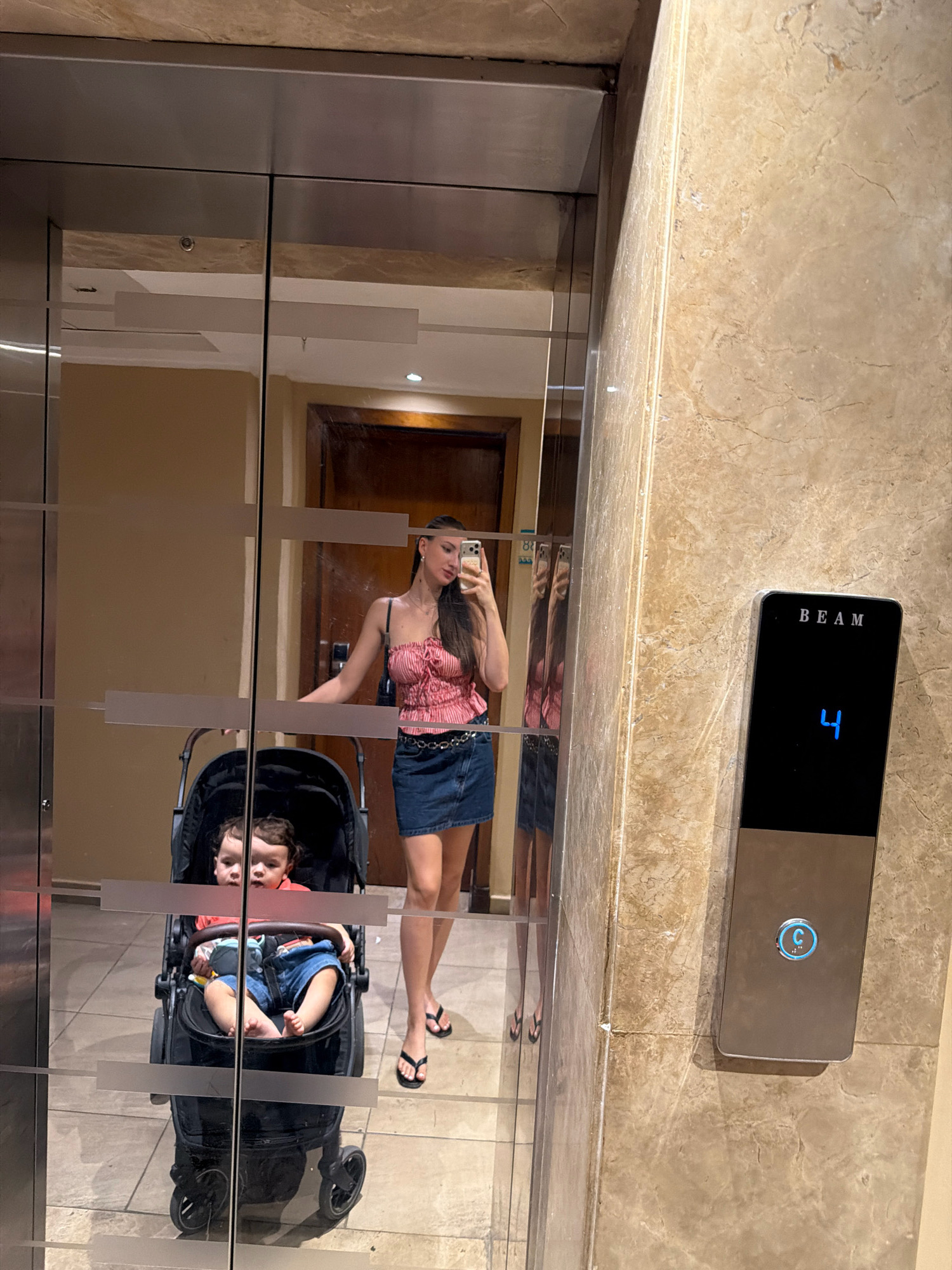 A young woman with dark hair, wearing a pink and white striped halter top, a denim skirt, and black flip-flops, is taking a mirror selfie in an elevator. She is holding a smartphone in her right hand. A baby is seated in a black stroller next to her, wearing a red shirt and denim shorts. The elevator has mirrored walls and marble accents. An elevator control panel with the number "4" illuminated in blue is visible on the right side of the image. The scene appears to be indoors, likely in a hotel or residential building. The location is Sharm El Sheikh, Egypt.