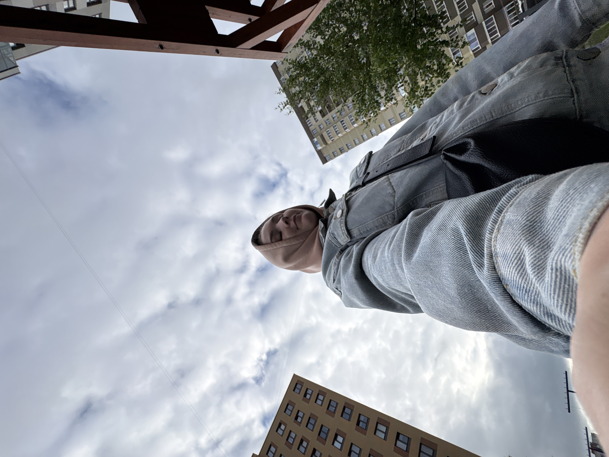 The image is a low-angle outdoor selfie featuring a person in an urban setting identified as Izhevsk, Russia. A female individual, appearing young to middle-aged, occupies the foreground, holding the camera with an extended arm. She wears a faded light blue denim jacket over a light brown or peach-colored hooded garment that covers her head. A black strap from a bag crosses her torso, and a black bag is visible at her side. Her eyes are partially closed, and her facial expression is neutral. The background features two multi-story residential buildings. On the left, a tall building with a yellowish-brown facade and numerous dark rectangular windows is prominent. On the right, another tall residential building with a lighter, possibly beige or light grey facade, visible windows, and some balconies, is partially obscured by a leafy green tree. A dark brown wooden structural element is also visible on the far right edge of the frame. The sky is overcast with scattered white and grey clouds, and two parallel thin dark lines, consistent with overhead power lines or cables, diagonally cross the upper central portion of the sky.