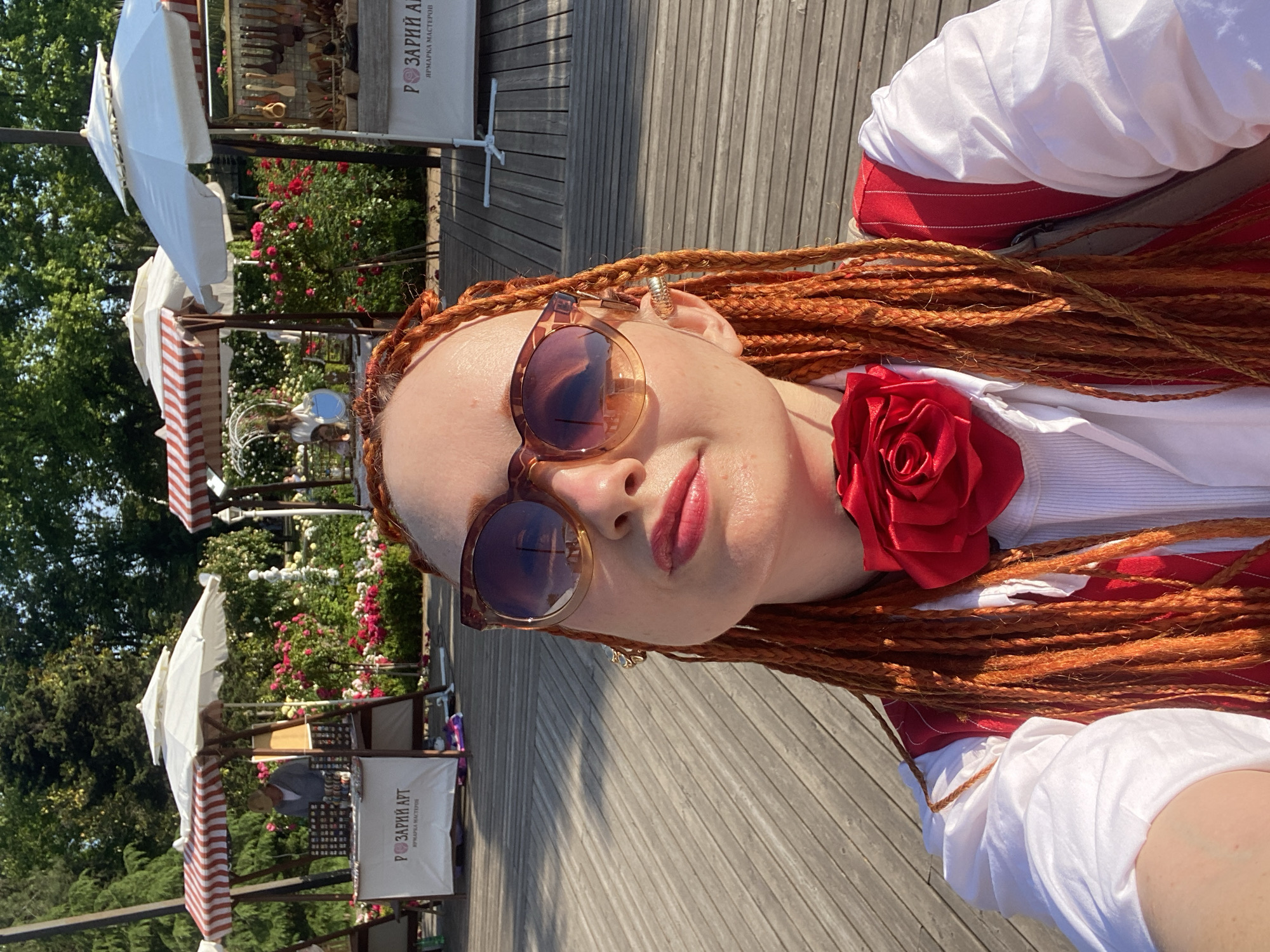 The image depicts a woman with bright orange braided hair, wearing oversized sunglasses, a white shirt, and a red vest adorned with a large red fabric rose at the collar. She is taking a selfie outdoors on a wooden boardwalk. In the background, there are market stalls with white umbrellas and red and white striped awnings, set up amidst lush greenery and vibrant pink and red roses. One stall displays wooden utensils. Another stall has a banner with Russian text, likely advertising "Rosarium Art" and "Craft Fair". In the distance, a person is visible at one of the stalls, and more people are partially visible near other stalls. The setting appears to be a park or an outdoor market, possibly a rose garden, in Sochi, Russia, given the mention of "Rosarium Art" which suggests a connection to roses. The lighting indicates a sunny day, with shadows cast on the boardwalk.