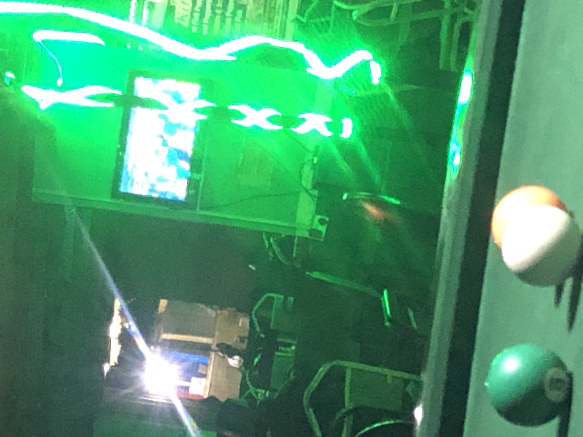 The image depicts a dimly lit indoor scene, possibly a bar or entertainment venue, with a prominent green neon light casting a strong glow throughout. In the foreground, two billiard balls are visible on a green felt surface. One is a solid green ball with a white number that is partially obscured. The other is a striped ball, white with an orange stripe, also with a partially visible number.
In the background, a television screen displays a bright, pixelated image. To the right of the television, a neon sign with white letters is illuminated against the green background. The sign appears to have a vertical element with horizontal crossbars, resembling a stylized ladder or abstract figure. The environment is filled with chairs and tables, suggesting a seating area. There are also reflections visible on the surfaces, indicating a polished or glass-like element in the scene. The overall atmosphere is dark, with the bright green light creating a dramatic and somewhat hazy effect. The location is identified as Unknown, Unknown.