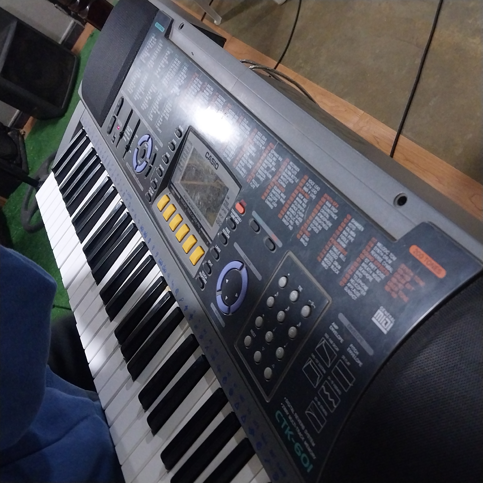The image shows a gray Casio CTK-601 keyboard. The keyboard has black and white keys, a display screen, numerous buttons, and built-in speakers. The top panel of the keyboard is labeled with various sound options, rhythms, and functions. Part of a person wearing a blue garment is visible on the left side of the keyboard. The keyboard rests on a wooden surface.