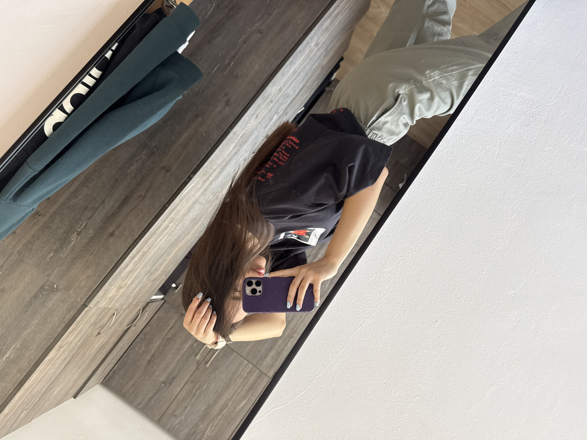 A person is reflected in a mirror, holding a purple phone. The individual is wearing a black t-shirt with some red writing on the back, and grey pants. The background shows a wardrobe with wooden paneling and a garment hanging inside, along with a white wall. The location cannot be determined to be Plovdiv, Bulgaria, based on the visual evidence provided.