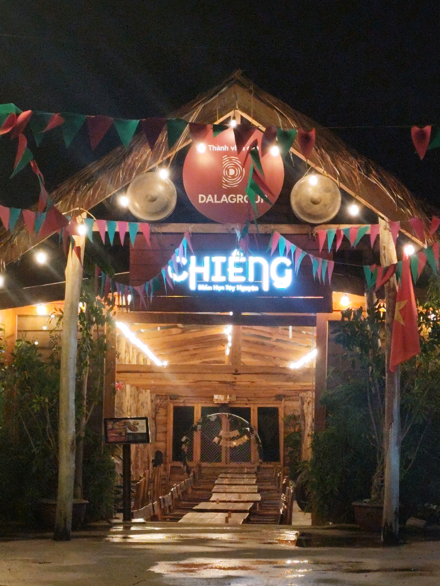 The image shows the entrance to a restaurant called "Chiêng" in Lâm Viên, Đà Lạt, Vietnam. The restaurant has a rustic, wooden construction with a thatched roof. Above the entrance is a neon sign that spells out the restaurant's name. Festive decorations are strung around the entrance, including pennant banners in pink, green, and red, along with hanging lights. A Vietnamese flag is visible on the right side. To the left of the entrance, a menu board stands on a post. The entryway has a set of wooden tables and chairs leading into the restaurant. Plant pots are on either side of the entrance. The ground in front of the restaurant appears to be wet, possibly from rain.