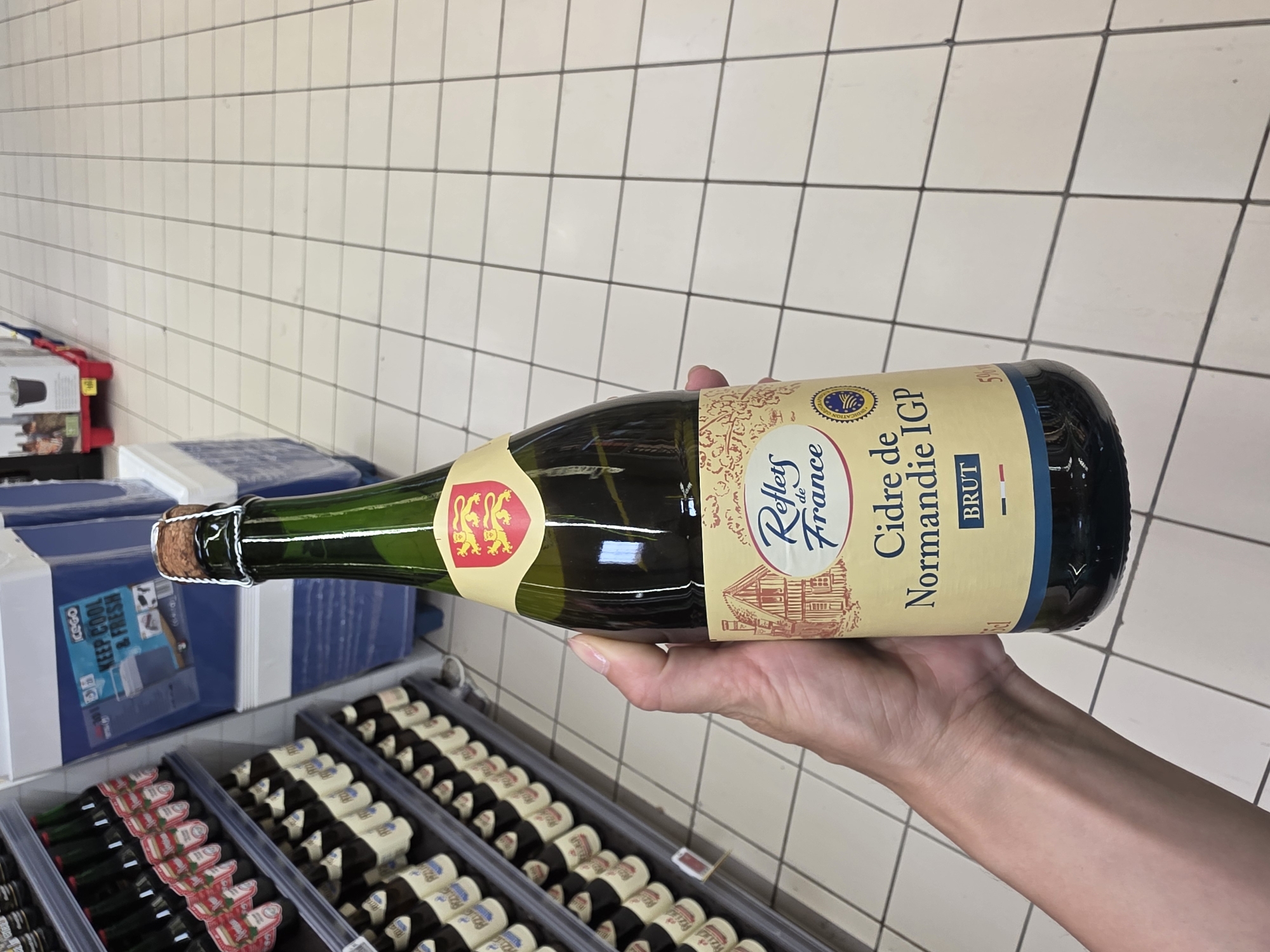 In the foreground of the image, a person's hand holds a green glass bottle of "Reflets de France Cidre de Normandie IGP Brut" which has a cork and wire closure. The bottle has a beige label with a drawing of a half-timbered house and a red and yellow shield with three lions. The background shows shelves stocked with more bottles and a display of blue and white coolers. The floor is tiled with a white grid pattern. This scene appears to be inside a supermarket.
