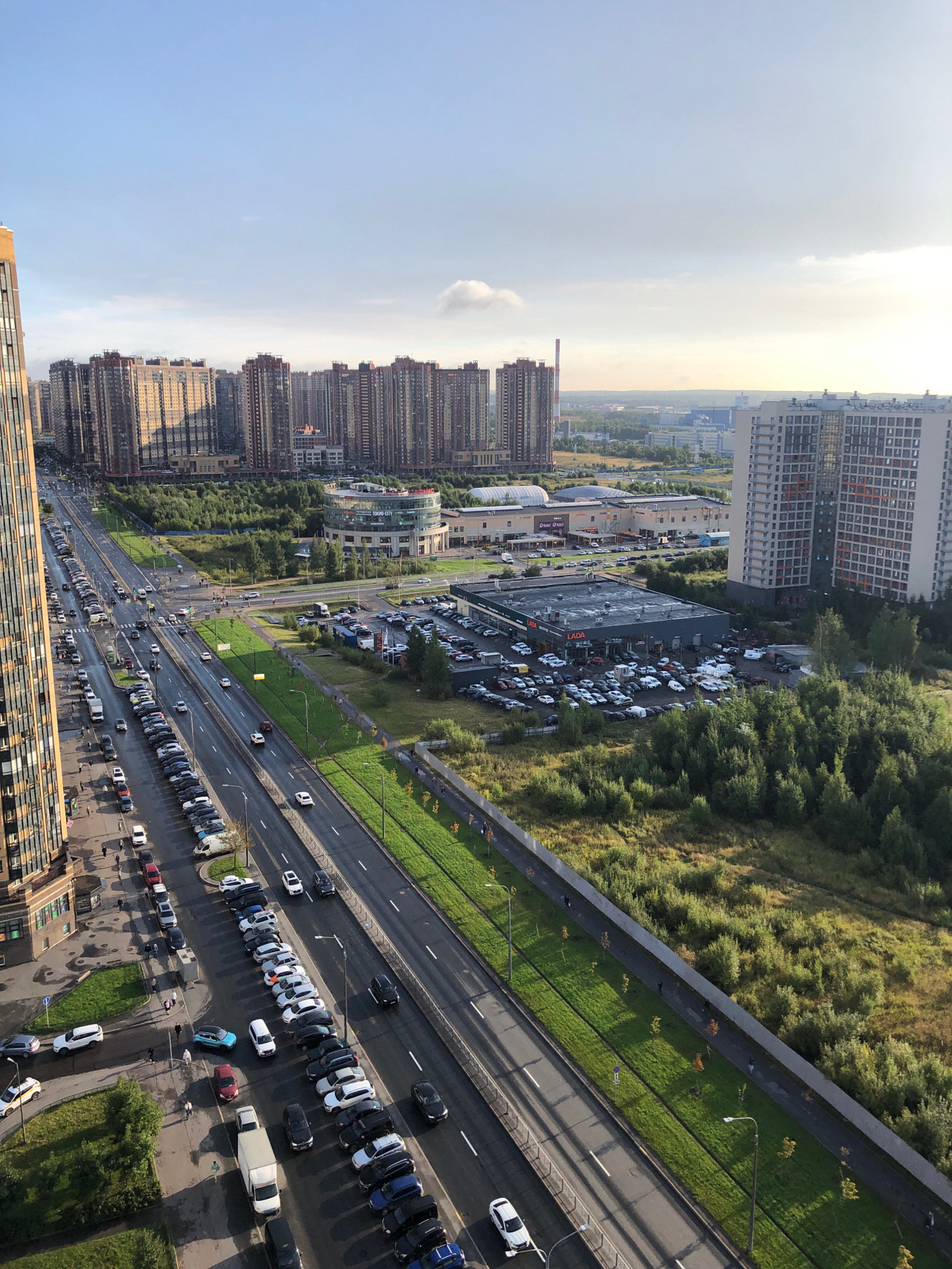 The image captures an aerial view of a multi-lane road in a city, likely St. Petersburg, Russia, on a clear day. The road is lined with parked cars on both sides, and traffic can be seen moving in both directions. Adjacent to the road, there is a green median strip with trees and lampposts. Beyond the median, there are residential buildings, a commercial building, and a wooded area. The sun is casting long shadows, indicating it is either early morning or late afternoon. Several people are visible walking along the sidewalk. A large number of cars are parked in a lot in front of a car dealership.