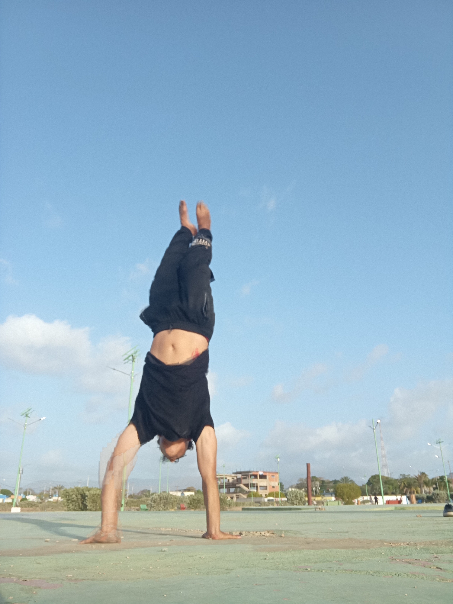 An outdoor shot captures a person performing a handstand on a light green, weathered ground surface. The individual is inverted, balancing on their hands with their head down and legs extended upwards. They are wearing a black t-shirt or tank top that has ridden up, revealing their midriff, and black pants with some indistinct white text or design on the right pant leg. Their feet are bare, and dark, curly hair hangs towards the ground.
The background features a clear, bright blue sky with scattered white clouds. In the mid-ground and distance, there are various elements suggesting an urban or semi-urban environment in Juan Griego, Venezuela. These include several modern-looking streetlights with green poles, low-rise buildings, sparse green bushes, and a few palm trees. A tall, slender brown structure, possibly a pole or monument, stands out in the mid-distance on the right. The ground beneath the person is a cracked, light green surface, possibly concrete or a faded sports court. The lighting suggests it is daytime, under clear weather conditions.