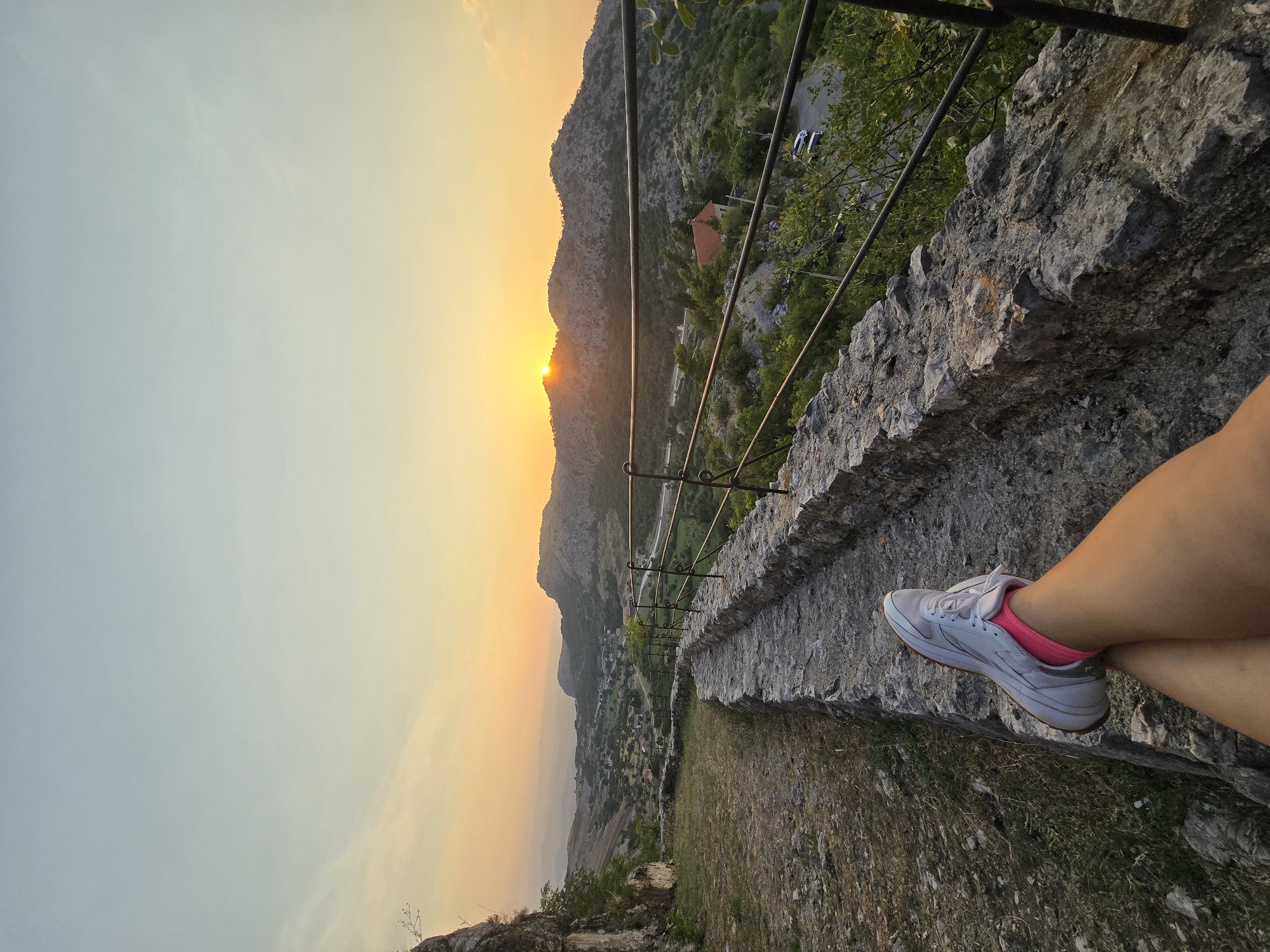 The image shows a first-person perspective of someone sitting on a stone wall overlooking a valley at sunset. The person's legs, clad in white sneakers and bright pink socks, are crossed in the foreground. The background features rolling hills covered in greenery, with a village nestled in the valley below. The sun is setting behind the hills, casting a warm orange glow across the sky and landscape. A metal railing runs along the stone wall, providing a boundary. The area appears to be an elevated viewpoint, possibly a historical site or a scenic overlook. The location is identified as Gornja Vrbica, Montenegro.
