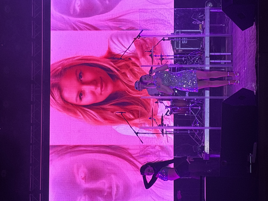 A woman in a glittering green dress sings on stage, with a large screen behind her displaying a close-up image of another woman with blonde hair. A dancer in a matching sequined top and black pants performs in the foreground. The stage is cluttered with microphone stands and equipment. The lighting is primarily pink and purple, creating a vibrant atmosphere.