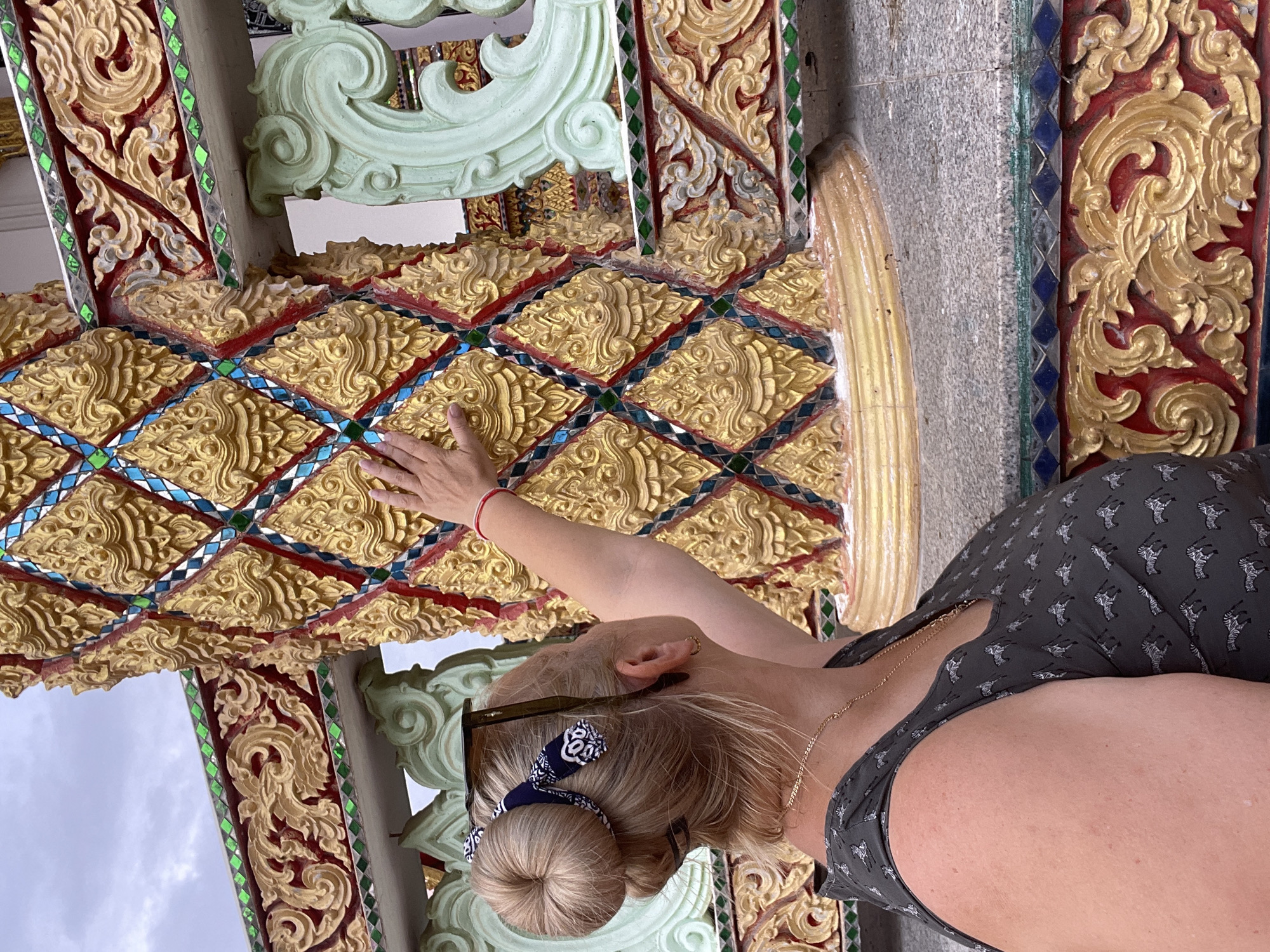 A woman with blonde hair styled in a bun and a dark grey top with a zebra pattern is touching a pillar decorated with gold, red, and blue mosaic tiles. She is wearing sunglasses on top of her head, a necklace, and a red bracelet on her left wrist. The pillar is part of an ornate architectural structure with intricate gold carvings and green decorative elements. The scene appears to be outdoors, possibly at a temple or a similar ornate building, with a cloudy sky in the background. The location is identified as Pattaya City, Thailand.