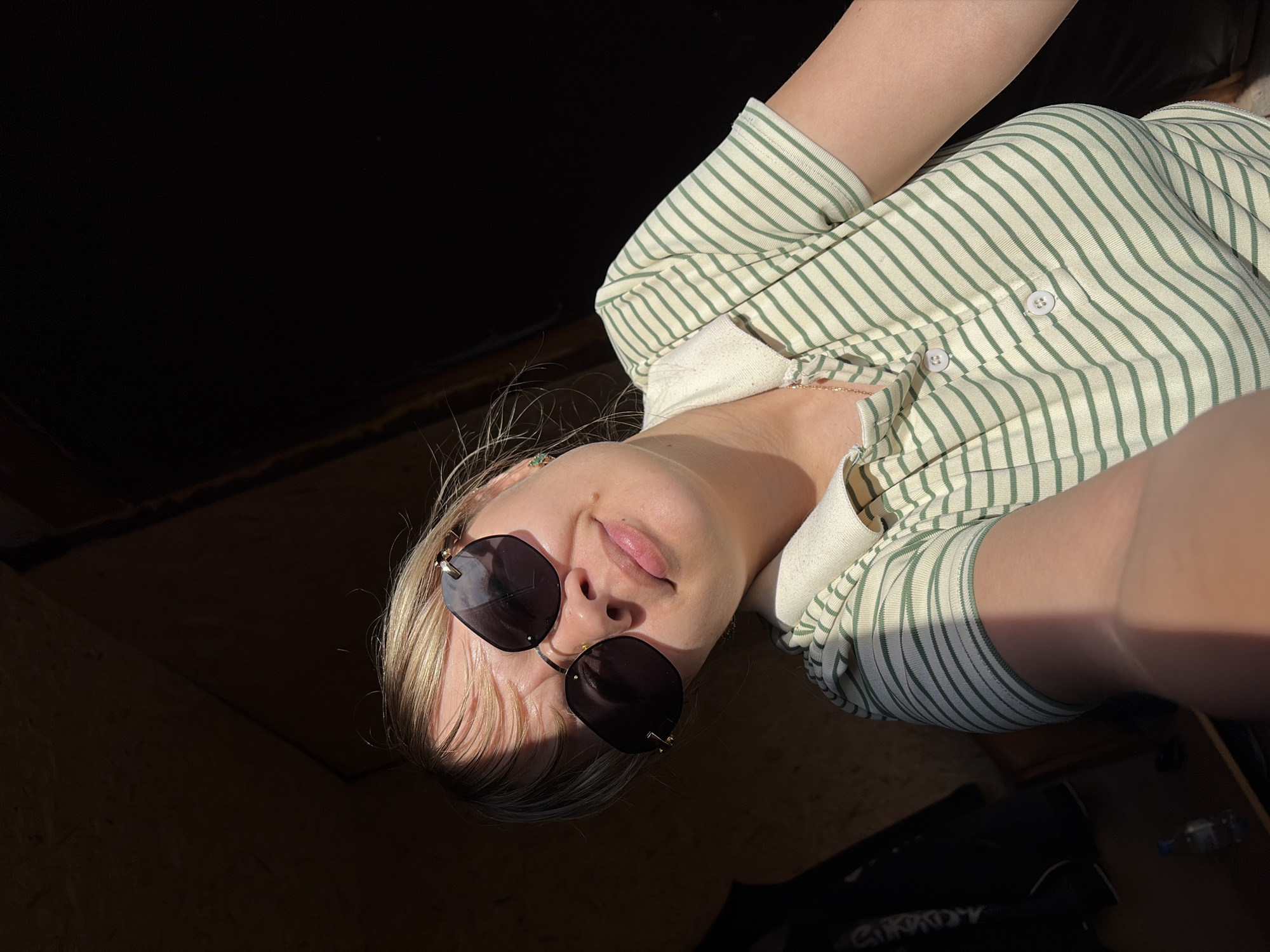 A person wearing a striped, button-up shirt and sunglasses is shown in a selfie-style shot. The background is dark, with areas of light and shadow. A portion of flooring and what appears to be a black bag are visible in the lower right corner. The location is not discernible as Чумна, Russia.