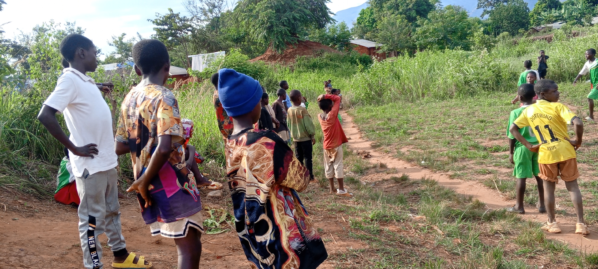 In Ngalula, Democratic Republic of the Congo, a group of children stand outdoors on a dirt path surrounded by green vegetation and trees. Some children wear colorful clothing while others wear sports jerseys, including a yellow shirt with the word "DORTMUND" and the number "11" printed on it. A few buildings are visible in the background, along with a hill.