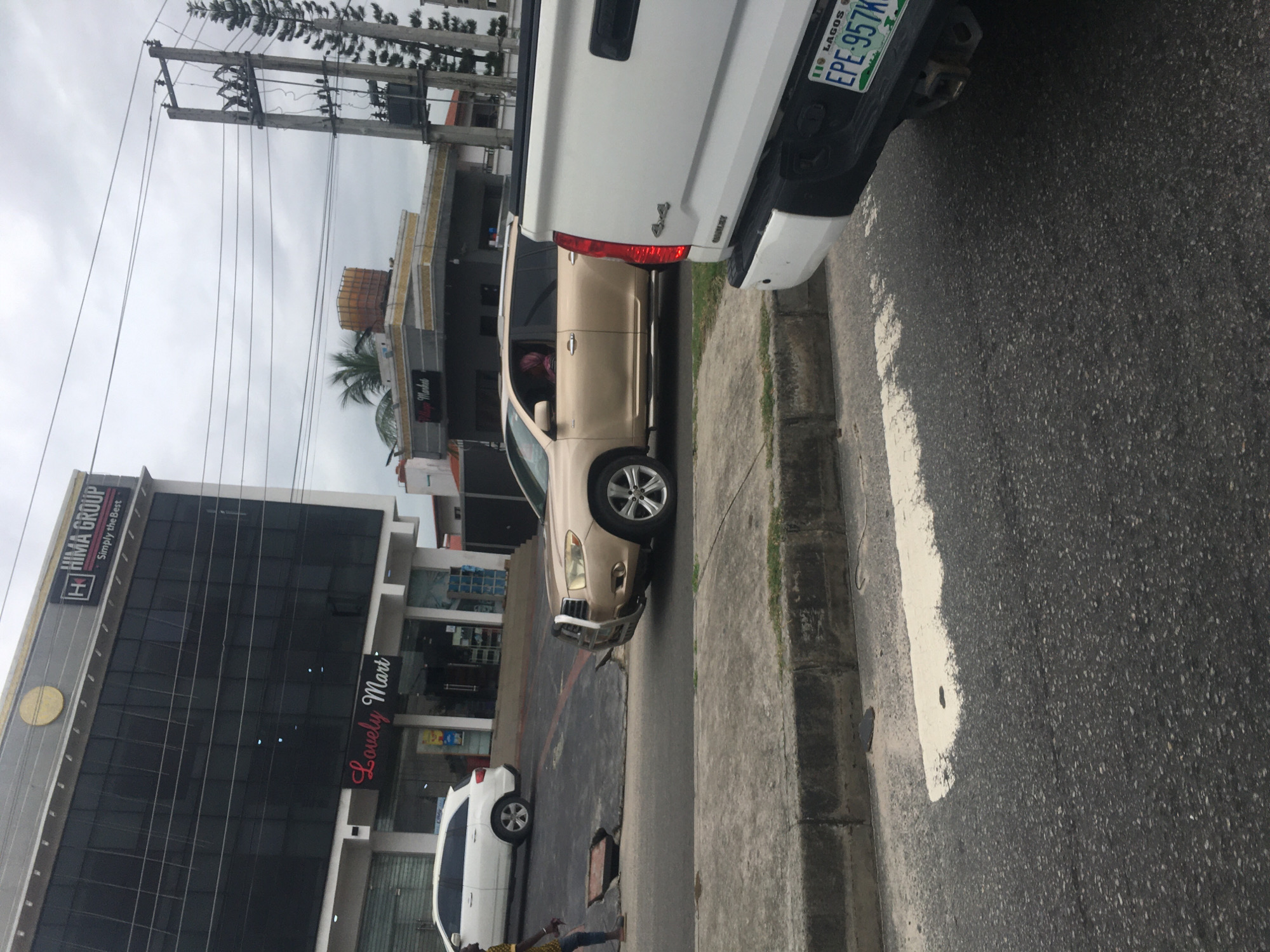 The image shows a street scene in Ado, Nigeria, with a white pickup truck partially visible on the right side of the frame, its license plate reading "IIO LAGOS EPE-957KT". A beige SUV is parked next to the truck, with its front driver's side tire resting on the curb. In the background, a multistory building is visible, displaying the signs "Hima Group" and "Lovely Mart". Another white car is parked further down the street. A pedestrian is visible at the bottom of the frame. Utility poles and overhead wires are also present. The overall environment suggests a commercial area within the city.