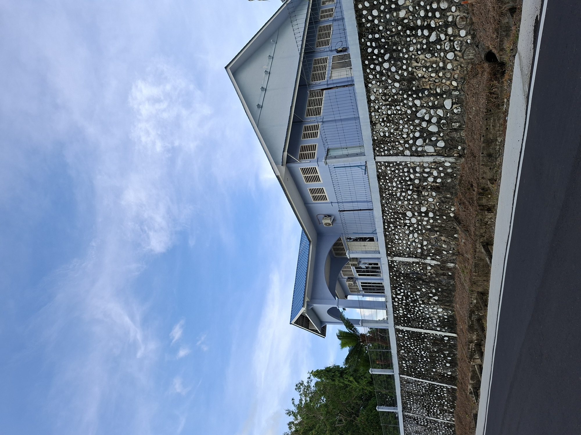 The image depicts a light blue, two-story building under a bright blue sky with scattered white clouds. The building's design includes a gabled roof, barred windows, and a partially covered porch area. A stone wall runs parallel to the building, separating it from a paved road with a white painted edge. Green trees are visible at the base of the building. The angle of the photograph is unusual, with the building positioned at a slant in the frame. The location is in Lhokseumawe, Indonesia.