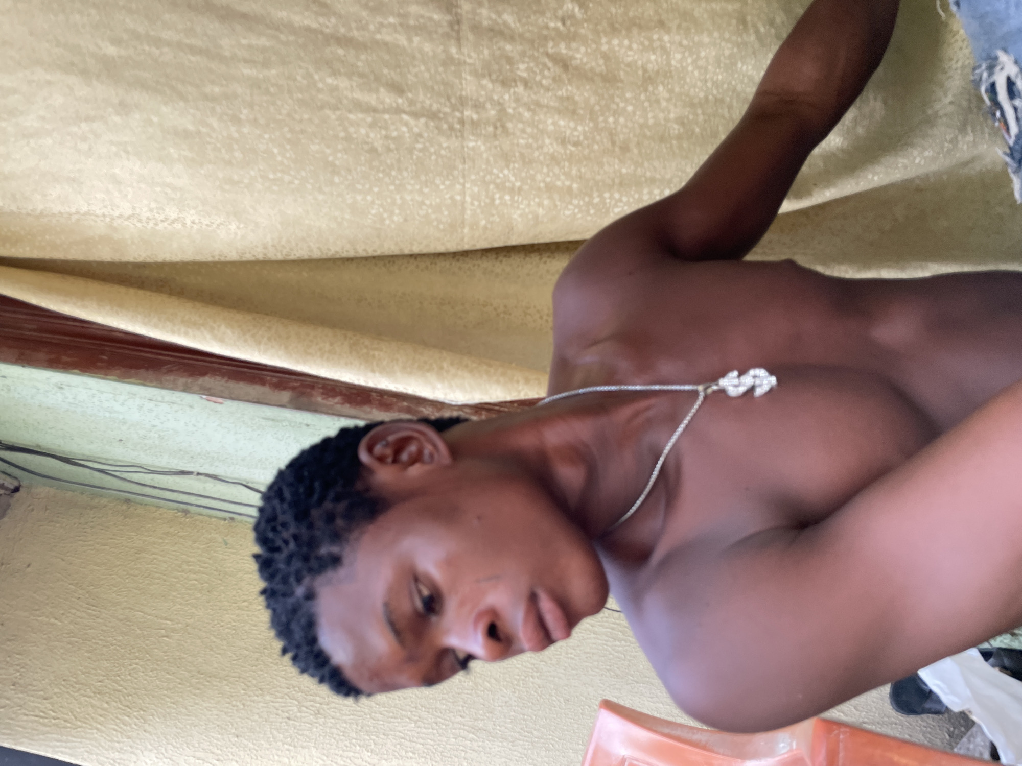The image displays a young adult male in a half-profile pose, facing left and looking downwards, captured in Ado, Nigeria. He has dark skin, short dark textured hair, and is bare-chested. A silver chain with a dollar sign pendant adorns his neck. A subtle linear mark is visible on his left cheek, below the eye.
The background on the left features a light yellowish-green textured wall, possibly stucco, with multiple dark electrical wires running vertically along its surface. A partially visible orange plastic chair or similar object is in the immediate left foreground. A reddish-brown vertical wooden frame or trim separates the wall from a light gold curtain on the right side of the frame. The curtain exhibits a subtle, repeating patterned texture and is draped in folds. A small section of ripped denim fabric is visible in the lower right corner, suggesting part of the subject's clothing or an item in the foreground. The lighting suggests natural, ambient illumination.