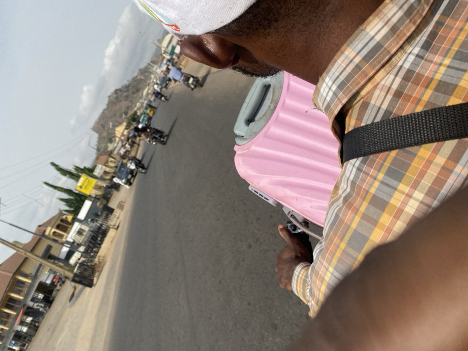 The image presents an outdoor, daytime scene in Nagazi, Nigeria, viewed from a first-person perspective, likely from the rear seat of a motorcycle or similar vehicle. In the immediate foreground, the back of an individual's head, right ear, and right shoulder are visible. The individual has dark skin, a short beard, and wears a white baseball cap with indistinguishable markings on the right side. They are attired in a plaid shirt patterned with shades of brown, yellow, and white. Their right hand is positioned as if gripping a handlebar or structural component of the vehicle.
Directly in front of this individual, or mounted to the vehicle, is a light pink, ribbed, hardshell case, possibly a suitcase or cooler, featuring a light grey handle visible at its top. A black strap, potentially from a bag or securing the case, crosses over the individual's right shoulder.
The scene extends onto a paved asphalt road, occupied by multiple motorcycles, some carrying passengers, and at least one white car. The vehicles appear to be in motion, traveling along the road.
To the left of the road, several buildings are visible, including a two-story structure with a light-colored facade and a dark roof. Further down the road, another building displays a yellow sign. Utility poles with overhead lines and scattered vegetation line the roadside. In the distant background, a prominent, rugged, rocky hill or mountain is silhouetted against a partly cloudy, bright blue sky, indicating clear weather conditions.
