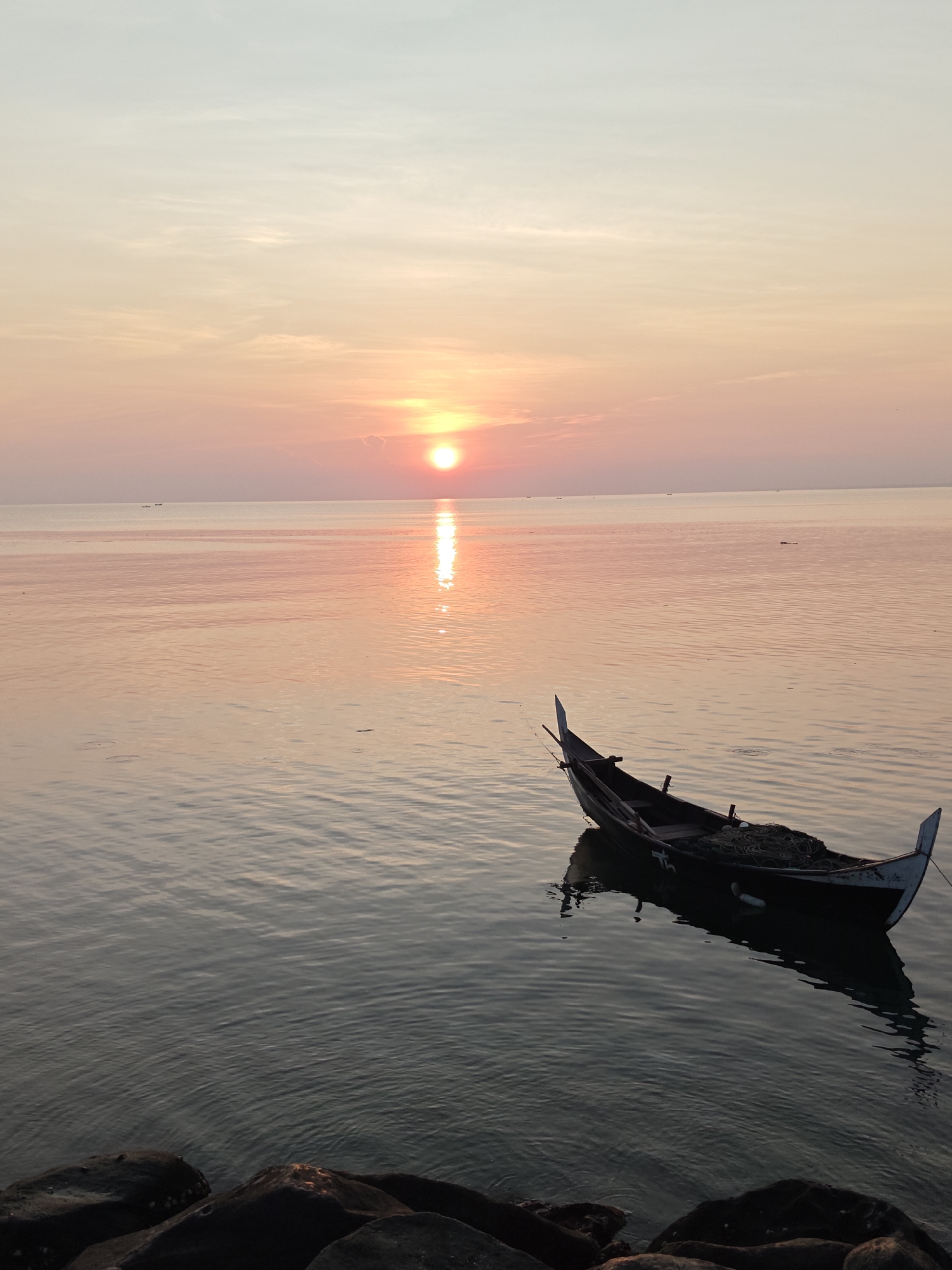 The image depicts a serene seascape at sunset. A traditional fishing boat is anchored in calm water. The sun is positioned near the horizon, casting a vibrant reflection on the water's surface. The sky is a gradient of soft pastel colors, transitioning from orange near the horizon to lighter hues above. Rocks are present in the foreground. The location is inferred to be in Lhokseumawe, Indonesia, based on the presence of the traditional fishing boat. There are no visible people or interactions.