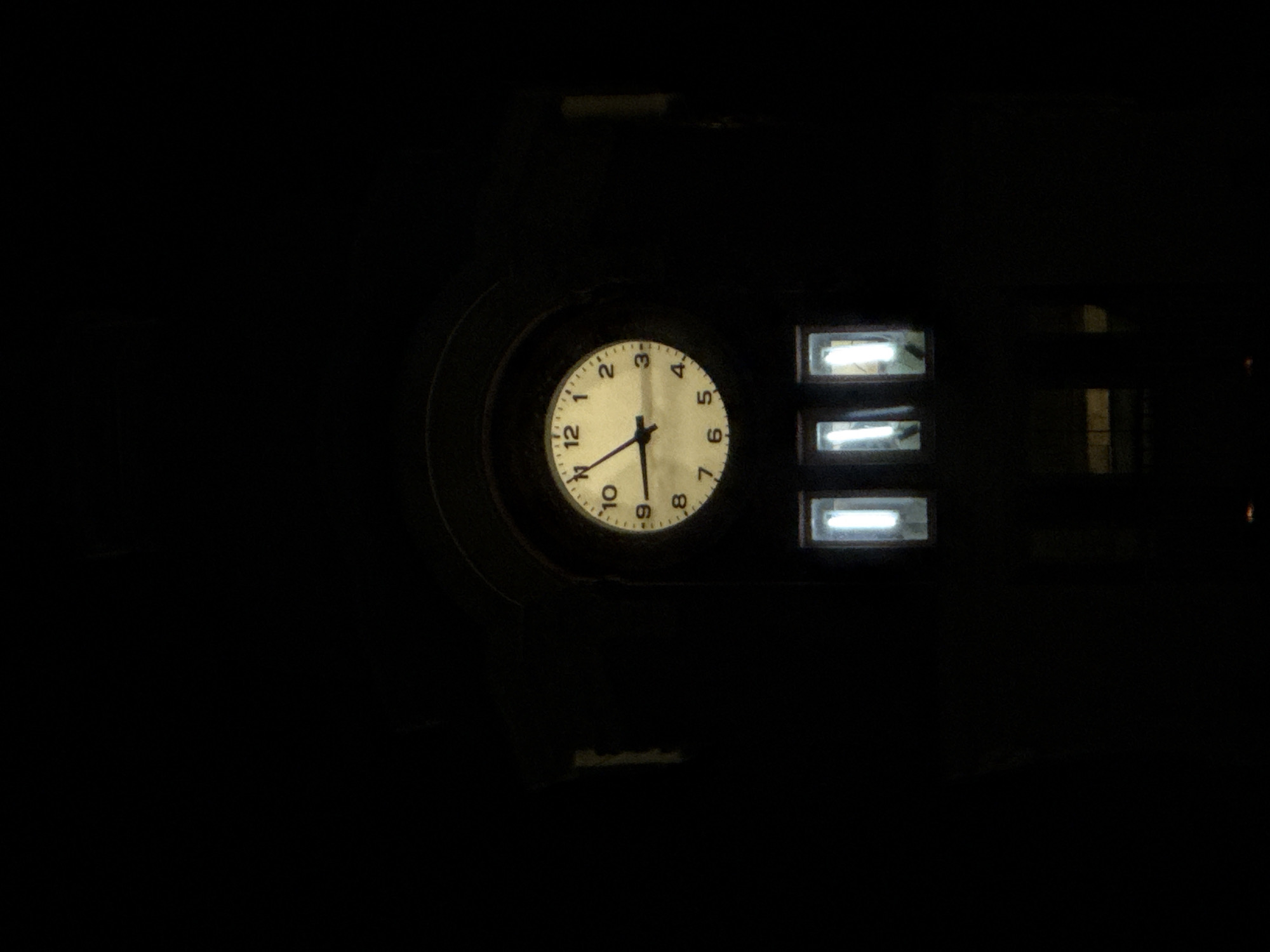An image of a clock mounted in a dark structure, likely a building, in Varna, Bulgaria. The clock has a white face with black numbers and hands indicating a time around 11:32. To the right of the clock are three horizontally oriented rectangular windows with bright, illuminated bars within. The setting appears to be nighttime, as evidenced by the darkness surrounding the objects and the brightness of the clock and lights. The surrounding structure is dark and mostly unidentifiable due to the low lighting.