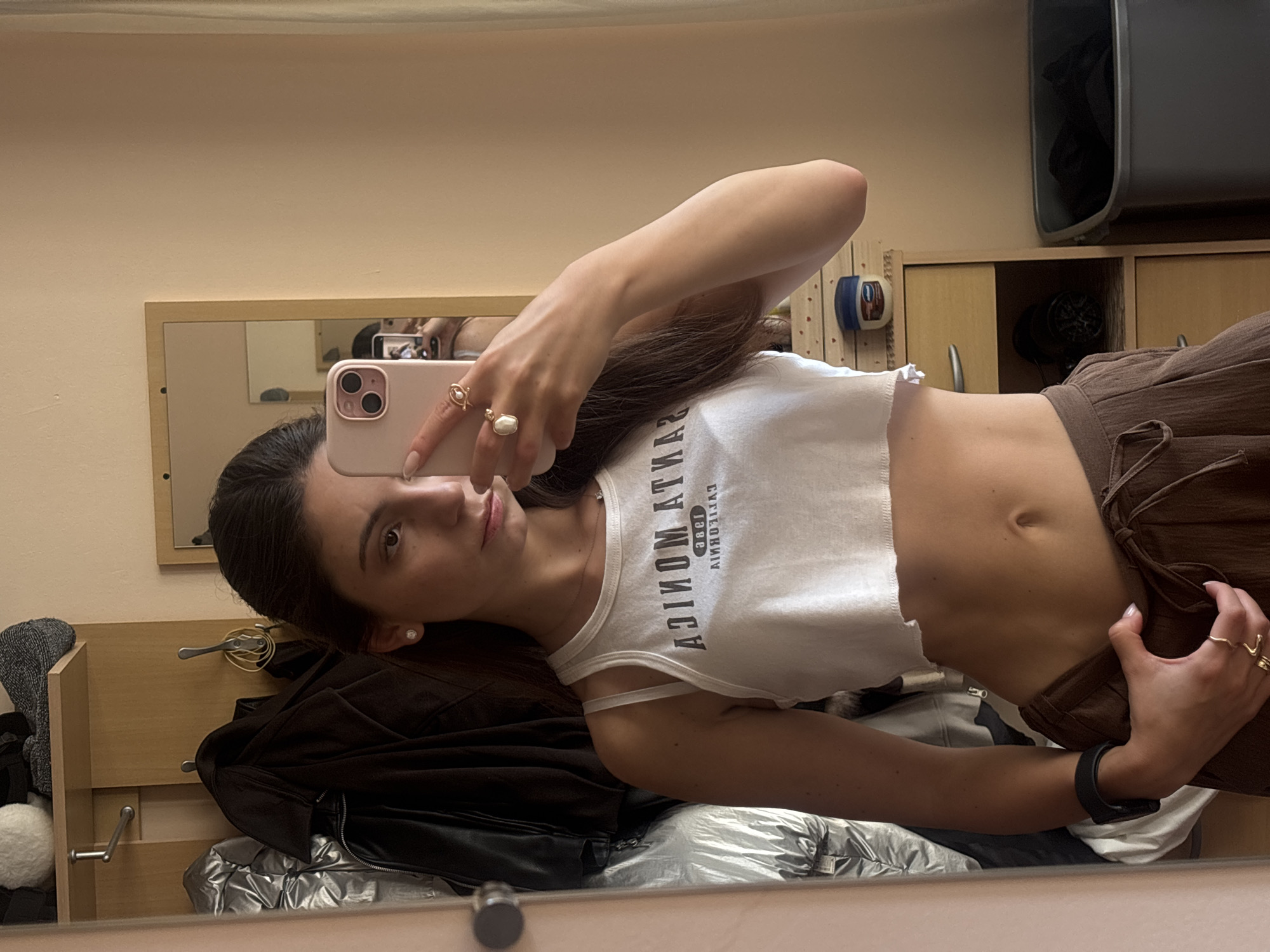 A young woman with dark hair pulled back takes a selfie in a mirror. She is wearing a white cropped tank top with "SANTA MONICA CALIFORNIA" printed on it and brown drawstring pants. She has on rings on her fingers and earrings. In the background, clothing hangs on hooks, and there is a dresser with a small jar of petroleum jelly and a trash can. This is likely a bedroom or closet area. There is no information in the image to indicate the city or country.