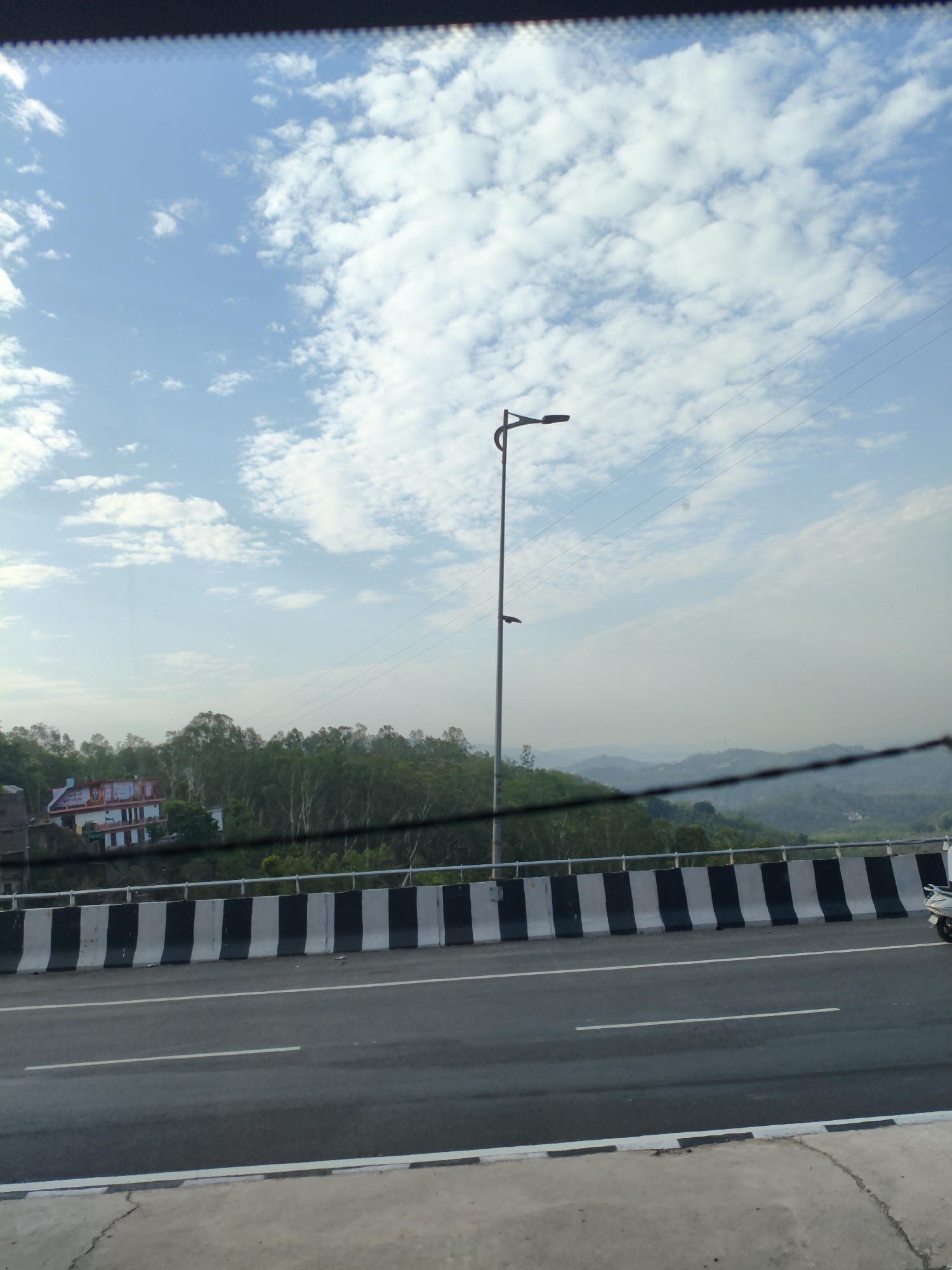 The image shows a highway in Bilaspur, India, on a cloudy day. The highway is divided into multiple lanes, and there is a concrete barrier with black and white stripes separating the lanes from the shoulder. A street light stands tall on the left side of the road. In the background, there are green hills covered with trees, and a few buildings are visible on the slopes. The sky is blue with scattered white clouds. The image is taken from a moving vehicle, as indicated by the blurred foreground and the slight motion blur in the image.