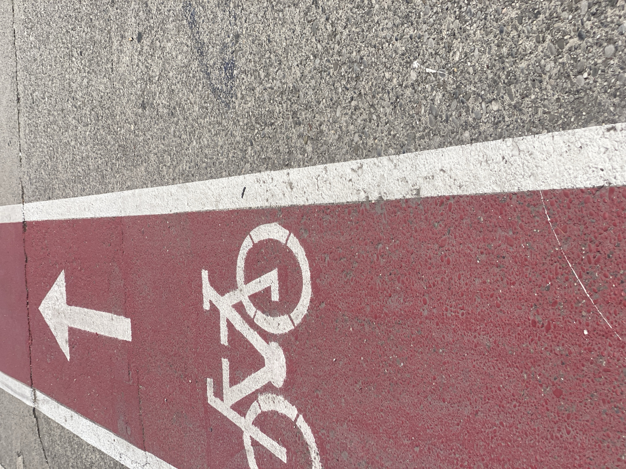 The image shows a close-up, high-angle view of a dedicated bike lane marked with a red surface and white symbols. A white arrow points upwards, indicating the direction of travel. Below the arrow, a white silhouette of a bicycle is painted. The bike lane is bordered by a white line and adjacent to a grey asphalt surface, likely a road or sidewalk. The asphalt shows signs of wear and contains small stones and cracks. The scene appears to be outdoors in a public urban space. The location is the pedestrian-bicycle path along the Morača river embankment in Podgorica, Montenegro.