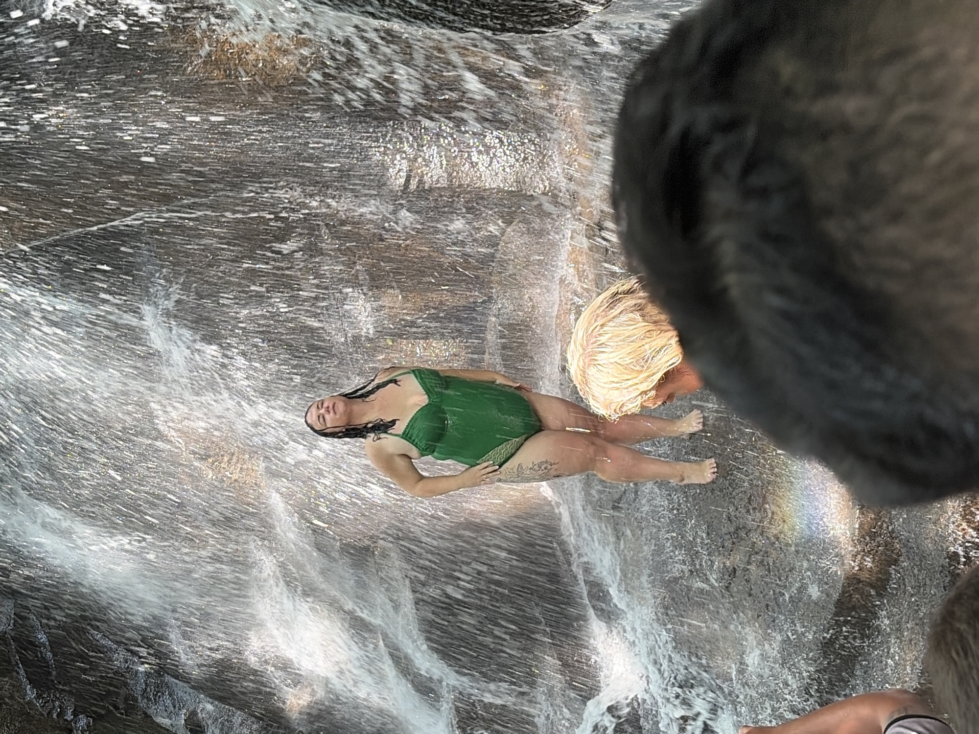 A woman in a green swimsuit stands under a waterfall. She has her eyes closed and her head tilted back. Her body is wet and her hair is dark and wet. She has tattoos on her left thigh and arm. To the right of her, a person with blonde hair is partially visible, and in the foreground, a person with dark hair is seen from behind. The waterfall cascades down rocks, and mist is visible in the air. The ground is wet and rocky, with some green vegetation visible. There is a small rainbow forming in the mist near the ground. The scene appears to be in a natural outdoor environment. The location in Gianyar, Indonesia is not identifiable from the image.