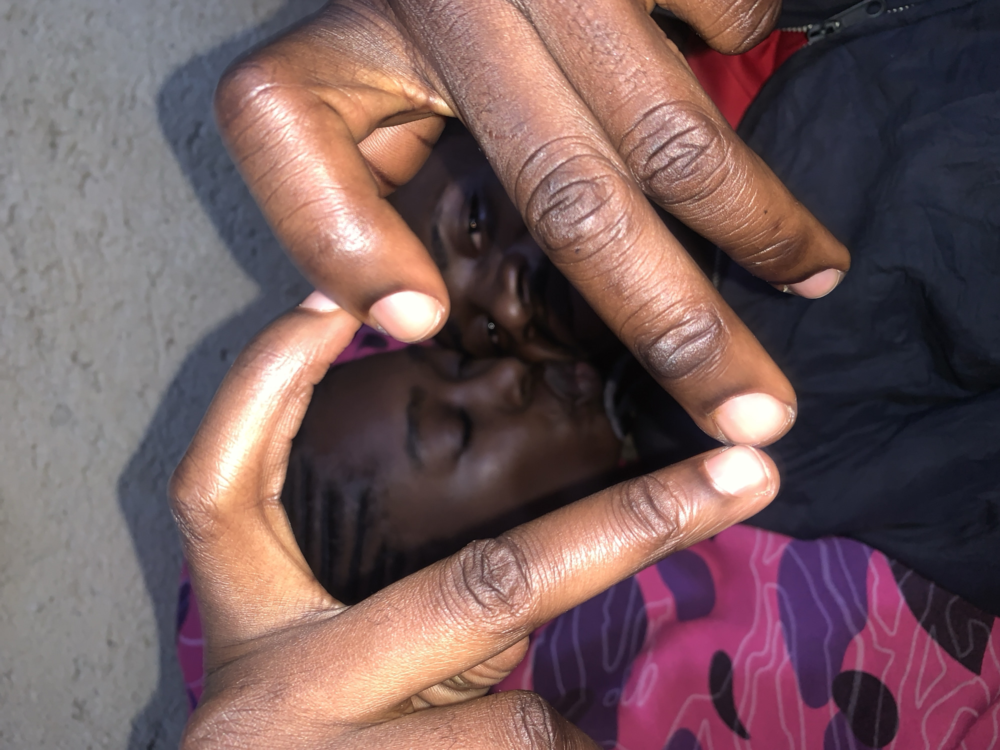 In Rafin Par, Nigeria, a close-up shot captures a loving moment between two individuals. Two dark-skinned hands are pressed together, forming a heart shape that frames the faces of a young man and woman. The woman, with braided hair, is seen in profile, her eyes closed and her lips pursed, suggesting a kiss. The man is partially visible behind her, his gaze directed towards the viewer. They are both dressed in dark clothing, and the woman is resting against a pink and purple patterned fabric. The background is a textured surface, possibly a wall or ground, with a shadow cast across it. The lighting is dim, emphasizing the intimacy of the scene.