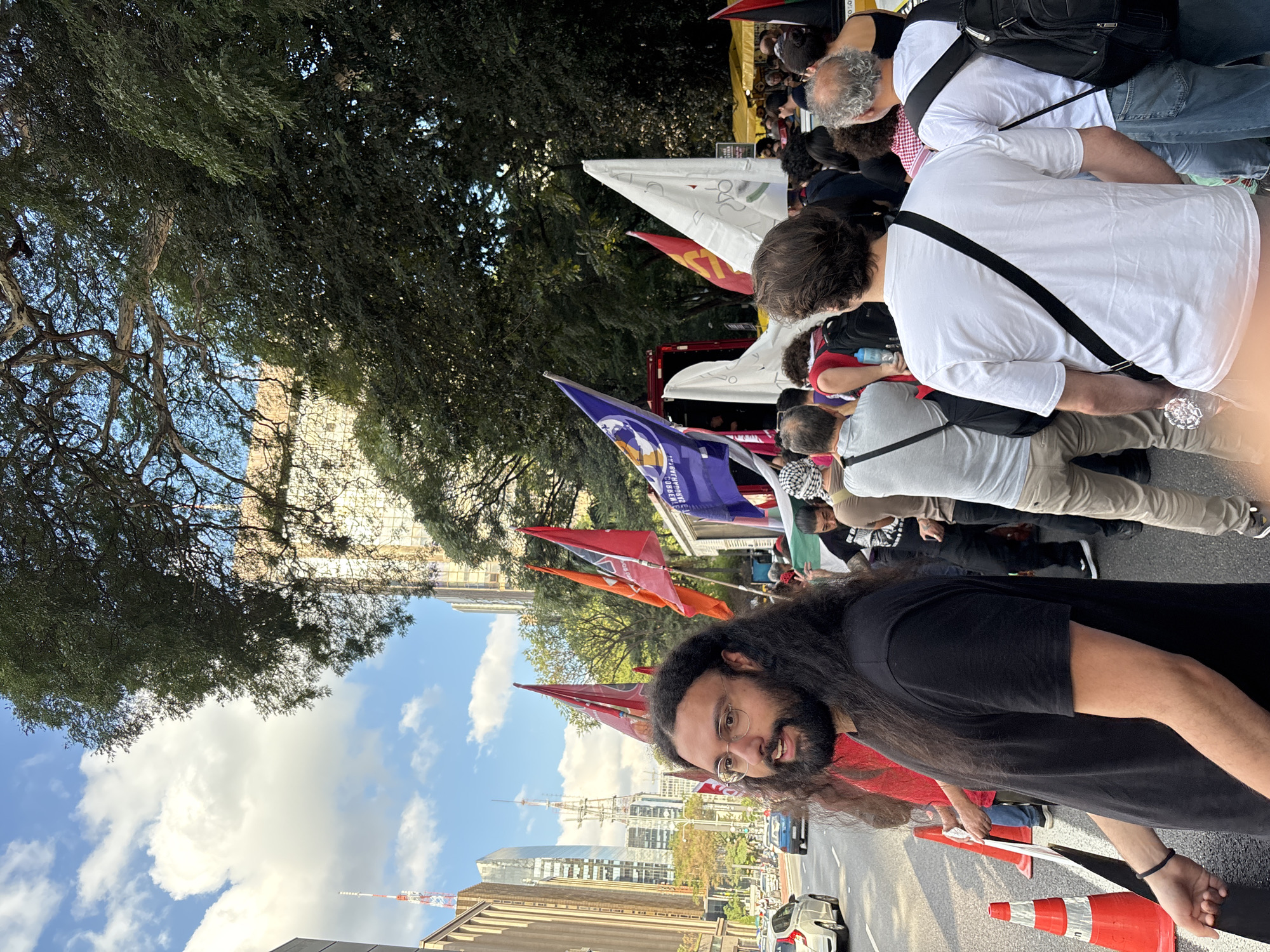 A medium-angle shot captures a political demonstration in São Paulo, Brazil, characterized by a crowded street lined with buildings and mature trees. In the foreground, a man with long dark hair and glasses looks directly at the camera, holding an orange traffic cone. Behind him, other participants hold red and other colored flags aloft. The crowd consists of people of various ages and ethnicities, some wearing white shirts and carrying bags. The architecture visible includes both shorter buildings and taller skyscrapers that punctuate the skyline, indicative of an urban landscape. The sky overhead is blue, dotted with scattered clouds. The angle and orientation of the image suggests it was taken while tilted or later rotated.