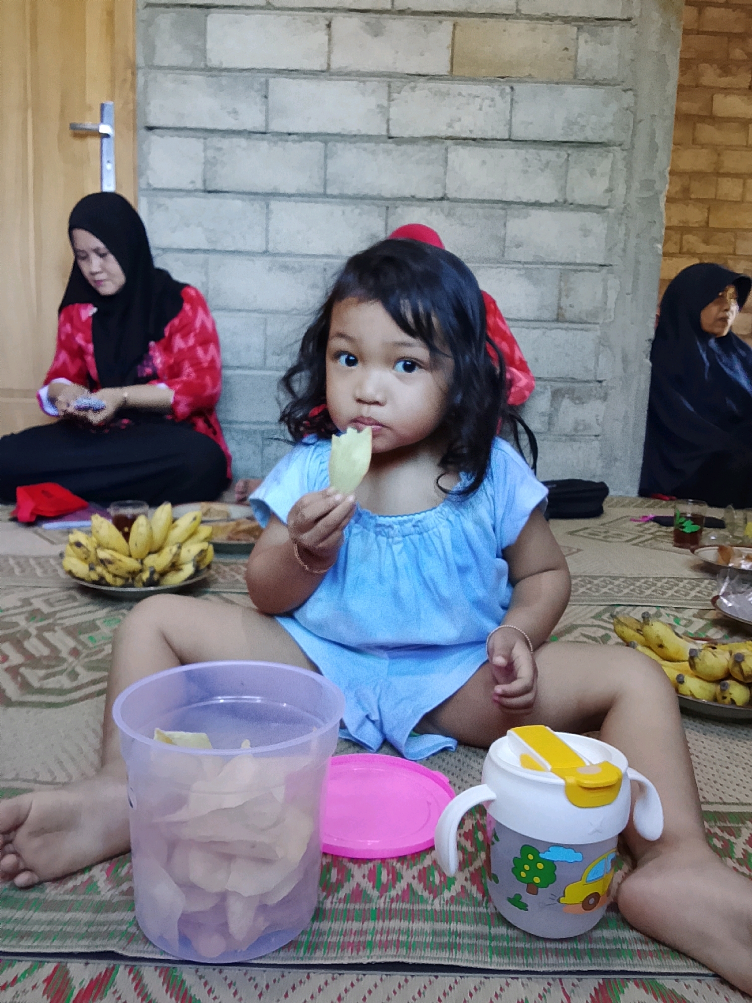 In Kradenan, Indonesia, a young girl with dark hair and brown eyes, wearing a light blue dress, sits on a woven mat, holding a light-colored snack. Her legs are spread wide, and her right hand is near her mouth. To her left is a clear plastic container filled with snacks and a pink lid. To her right, a sippy cup with a yellow lid and cartoon graphics is visible. In the background, a woman wearing a black hijab and a red and black patterned shirt sits on the mat, looking down at something in her hands. Behind her is a wall made of light-colored concrete blocks. To the far right, another person in a black hijab is partially visible. In front of the woman in red, a plate of bananas is displayed. The scene appears to be an informal gathering or meal.