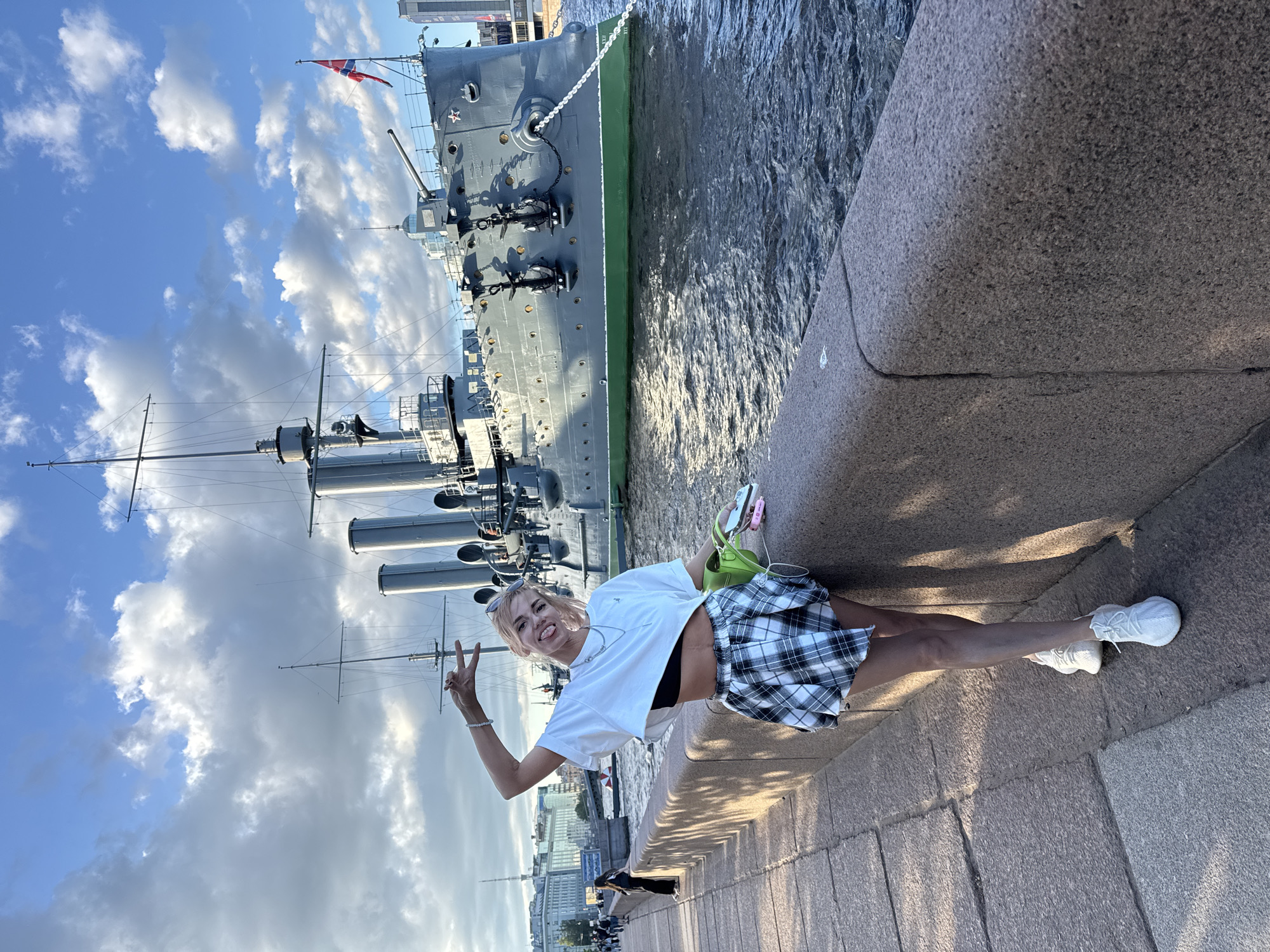 A young woman with blonde hair styled in a ponytail, wearing a white crop top, a black bralette, and a black and white plaid pleated skirt, stands on a granite embankment in Saint Petersburg, Russia. She is making a peace sign with her right hand and has her left arm bent with her hand resting on her hip. A lime green bag is tucked under her arm, and she holds a pink object in her left hand. Behind her, a large, gray warship, identified as the Aurora Cruiser, is docked in the Neva River. The ship has several smokestacks and is flying the Russian flag. The sky above is partly cloudy with scattered white clouds against a blue sky. The embankment is paved with large granite blocks, and a low stone wall separates it from the river. In the background, other people can be seen walking along the embankment, and buildings line the far bank of the river. The lighting suggests it is late afternoon or early evening.