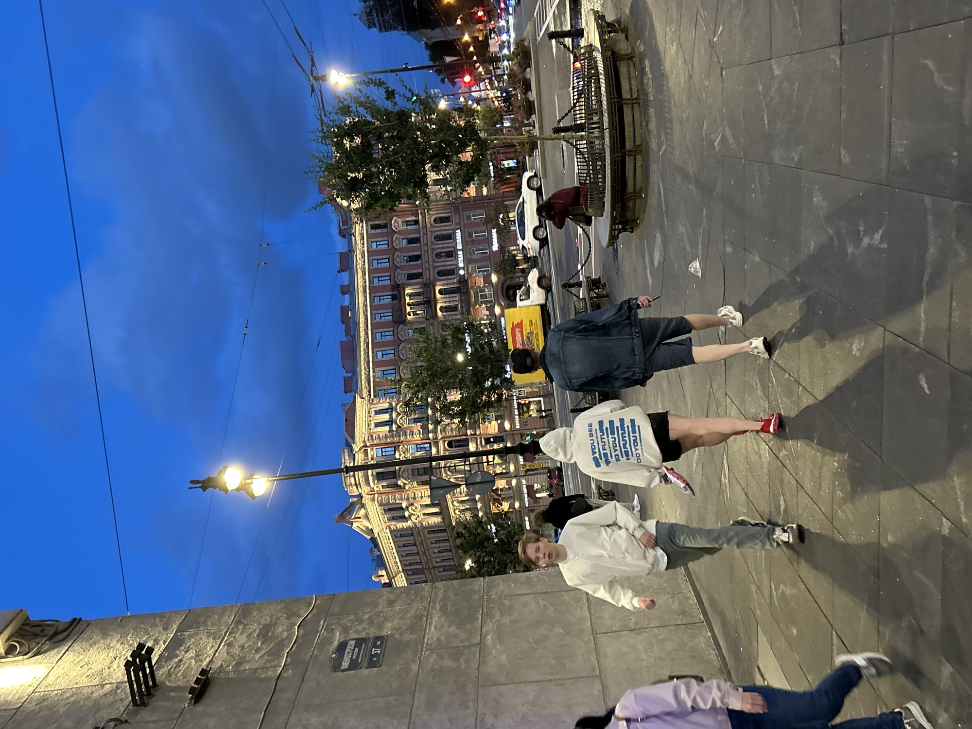 A wide-angle, outdoor shot captures a city scene at dusk. The sky is a deep blue, with some scattered clouds. The foreground features a sidewalk paved with dark rectangular tiles, casting long shadows from the figures walking on it. To the left, a large, textured concrete wall with visible fixtures and a street sign is prominent.
In the middle ground, several people are walking. Closest to the viewer, a young person in a light-colored jacket and grey pants walks towards the left. Behind them, two individuals walk away from the camera; one wears a white hooded sweatshirt with blue text and black shorts, and the other wears a dark denim jacket and dark shorts. Further back, other pedestrians and vehicles are visible on a street.
The background is dominated by ornate, multi-story buildings with illuminated windows, suggesting a commercial or residential area. A street lamp with two globes illuminates the scene. Several trees are also present, and a circular planter with a bench is situated on the street. Traffic lights and street lights indicate active city life. The overall impression is of a busy urban environment at the end of the day.