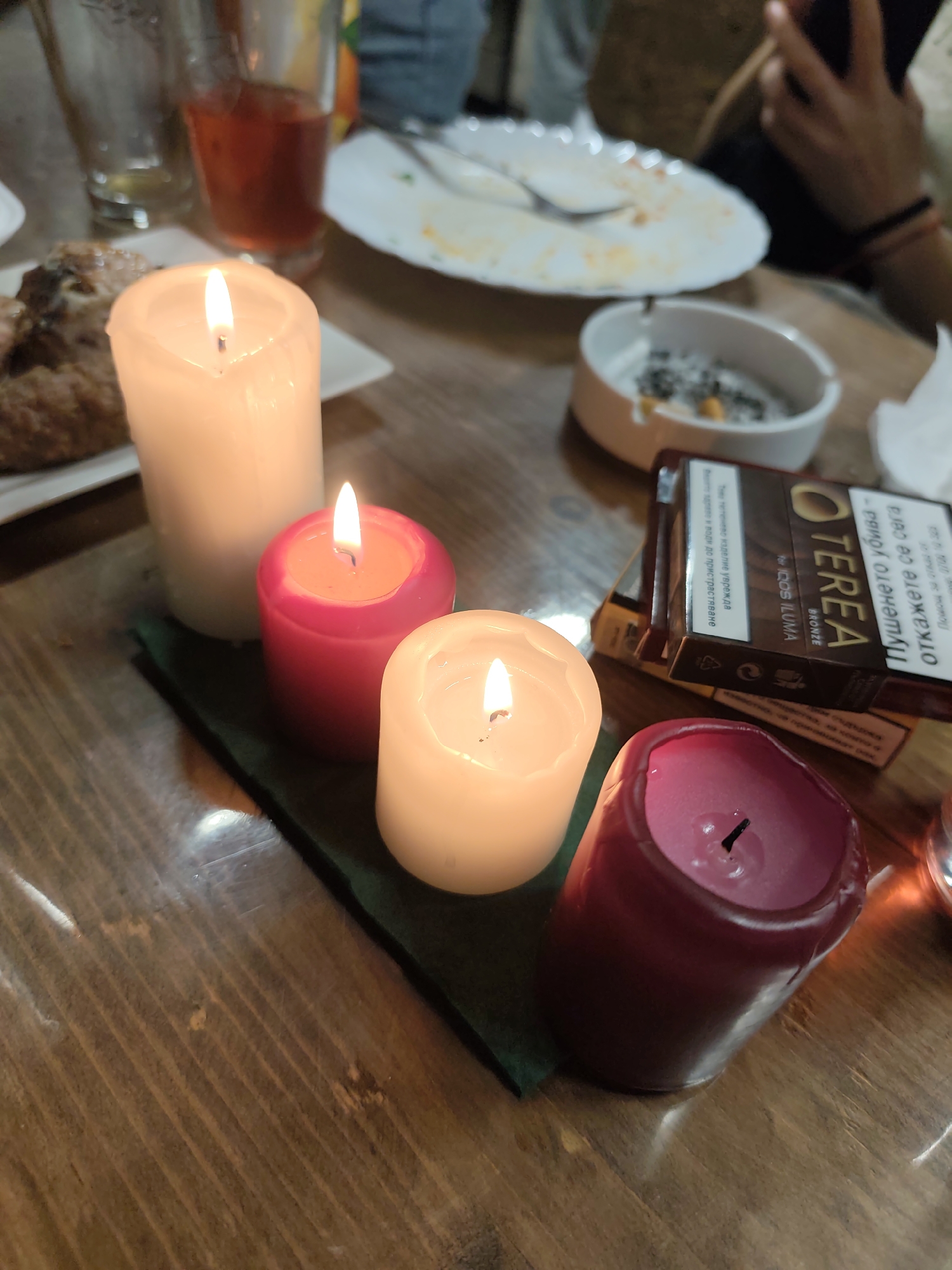 A lit candle arrangement sits on a wooden table. The arrangement consists of four candles of varying colors and sizes, placed on a dark green surface. The candles are white, pink, and red. Two are lit.
In the background, there is a white ashtray filled with cigarette butts, a pack of TEREA bronze cigarettes, plates with remnants of food, and glassware containing a reddish liquid. People are seated at the table, their arms and clothing visible in the periphery.
The image was most likely taken indoors in Mosomishte, Bulgaria.