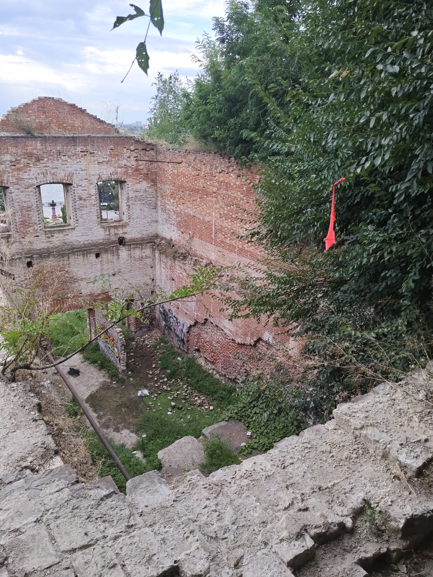 The image depicts the ruins of a brick building with large openings where windows once were, overlooking a body of water. The building is in a state of disrepair, with exposed brickwork and crumbling sections. Overgrown vegetation, including trees and bushes, surrounds the structure, with some branches reaching into the building's interior. A bright orange piece of fabric is tied to a branch of a tree, hanging prominently near the building. Graffiti is visible on some of the interior walls. The foreground shows rough concrete steps leading down towards the scene. The sky above is overcast.