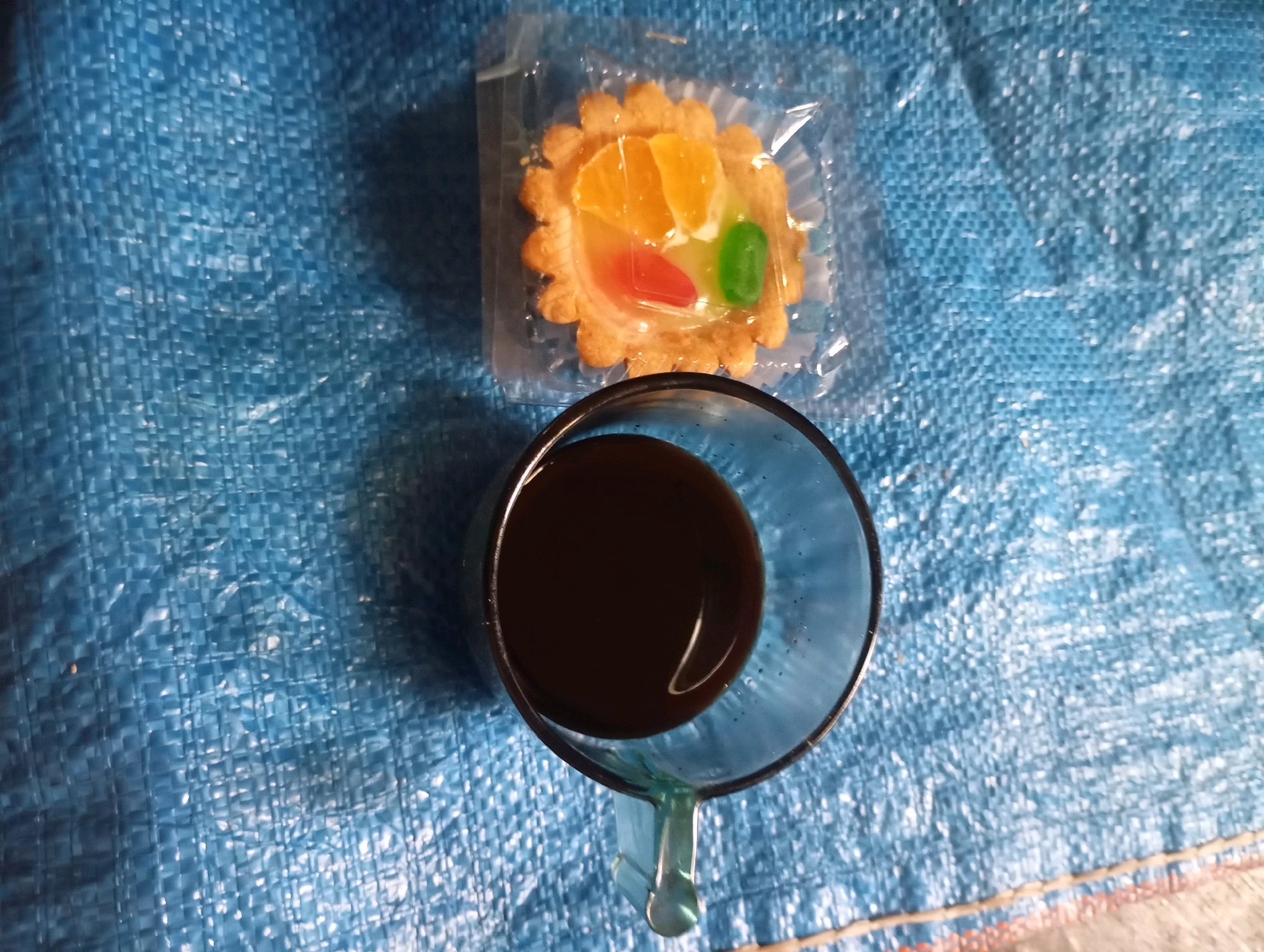 A fruit tart sits in clear plastic packaging next to a glass of dark liquid, possibly coffee, on a blue, textured surface that appears to be a woven tarp. The fruit tart has a scalloped crust and is topped with slices of orange and two pieces of colorful jelly, one red and one green. The glass is partially filled with the dark liquid, and its handle is visible at the bottom. The scene is lit from above, casting shadows. The location is in the city of Sumberjati, Indonesia.