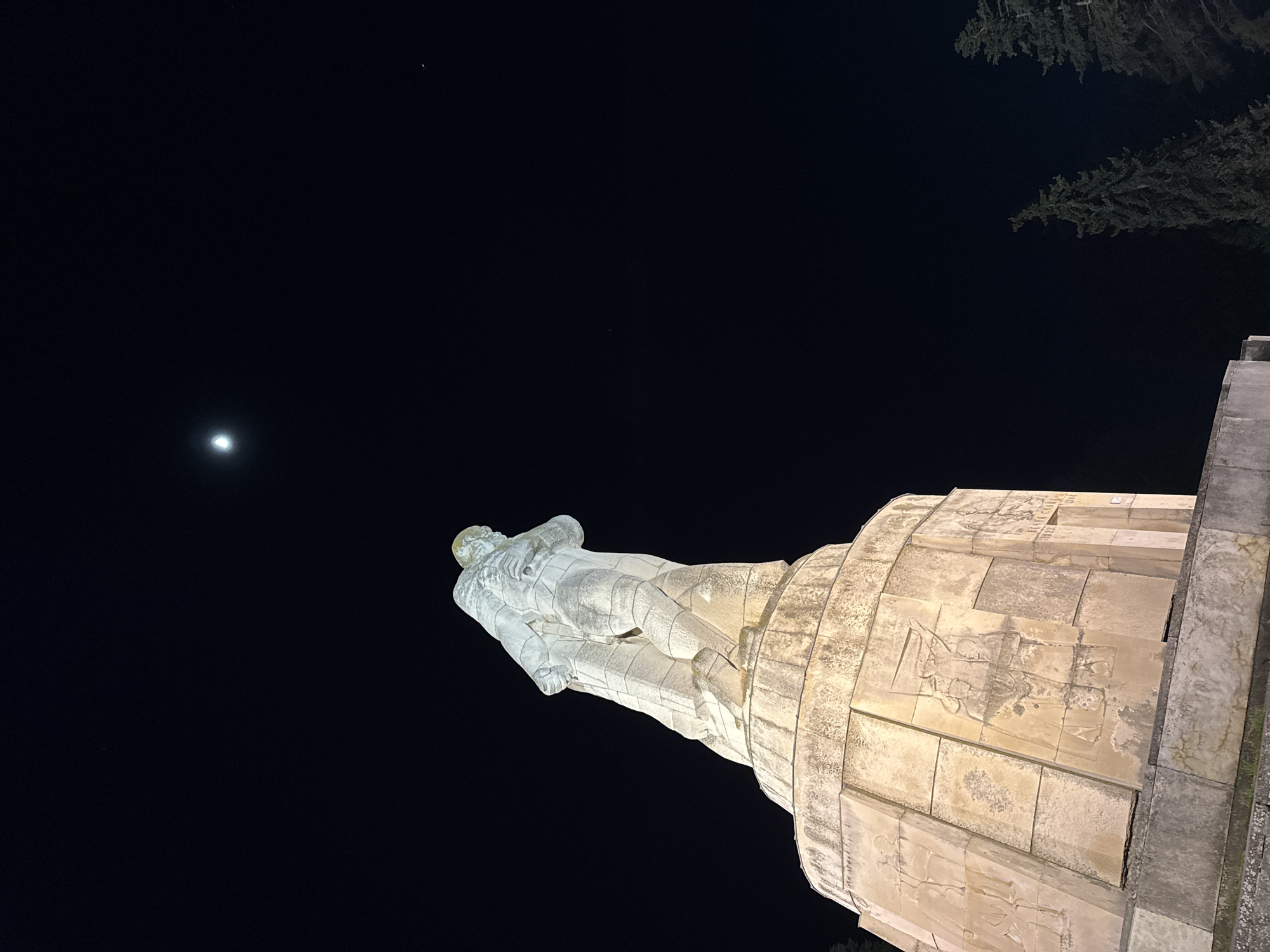 The image displays a large stone monument at night, dramatically silhouetted against a deep black sky. In the foreground, a significant statue of a male figure, likely a soldier, reclines horizontally on a substantial, circular stone pedestal. The figure, sculpted from light-colored, weathered stone, is illuminated from below, accentuating its form and details.
The pedestal is composed of segmented stone blocks, rising in several tiers, and is adorned with numerous bas-relief carvings. These carvings depict human figures, potentially representing historical or military scenes, though precise details are obscured by the distance and lighting. On a band above the bas-reliefs, carved text is partially visible, notably including the years "1923" and "1944" prominently, alongside Cyrillic letters that form parts of phrases like "СЛАВА НА" (GLORY TO).
The expansive night sky occupies a large portion of the frame. A bright, crescent-shaped moon or similar celestial body is visible towards the mid-left. A much smaller, fainter point of light, possibly a star or distant planet, is discernible in the upper-right sky. Dark, blurred outlines of tree branches extend into the frame from the top right corner. The photograph is taken from a low-angle perspective, emphasizing the monument's imposing size and its contrast with the dark night. The scene evokes a solemn and historical atmosphere, fitting for a monument in Varna, Bulgaria.