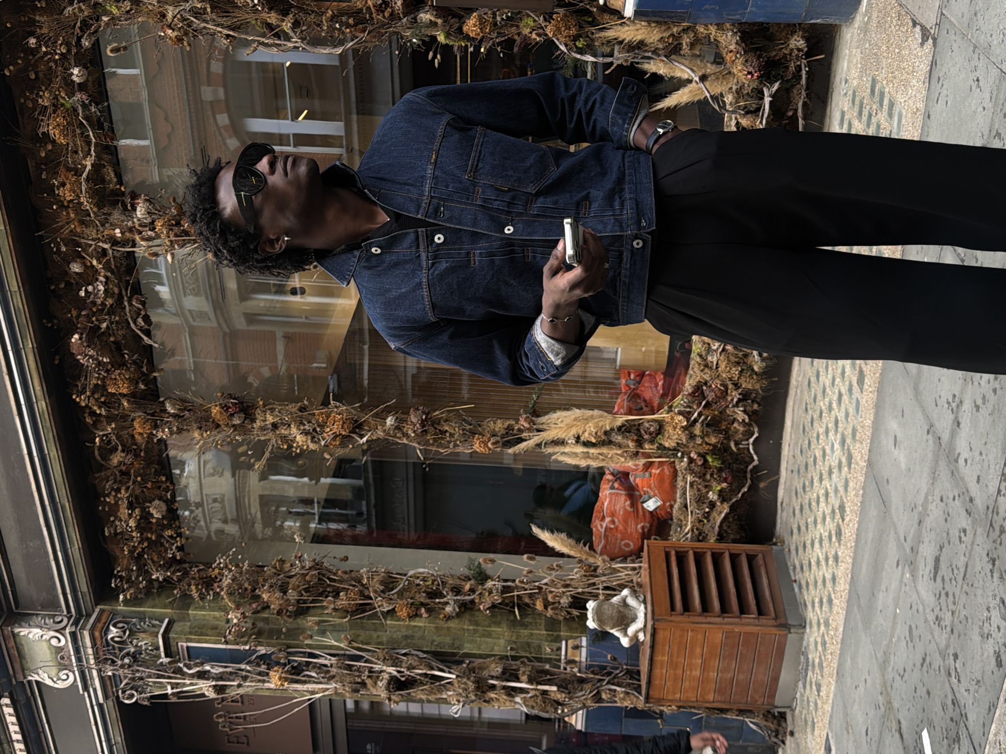 A dark-skinned male stands on a city street in front of a shop window. He wears sunglasses, a dark denim jacket, and black trousers. He holds a mobile phone in his right hand and his left hand is in his pocket. In the shop window are dried floral arrangements, and some orange bags. To the left of the man is a wooden cabinet with a frog statue on top. The street is paved with stone tiles, some of which are wet. The location is likely London, United Kingdom given the architecture visible in the shop window.