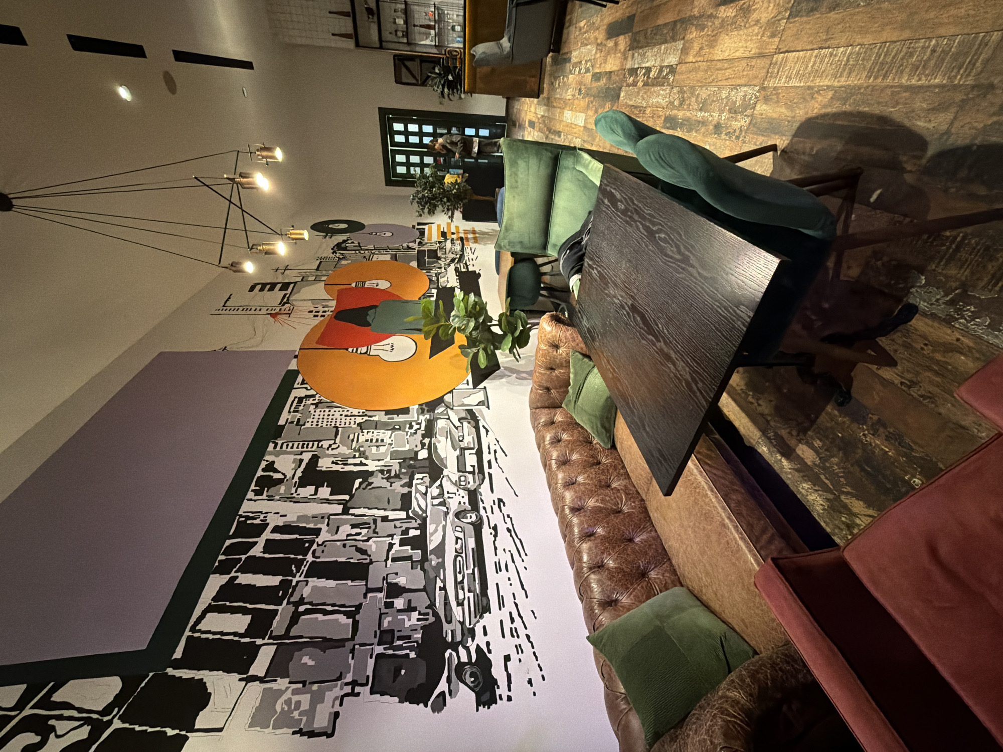 The image depicts a restaurant interior, likely in Tbilisi, Georgia, based on the design elements and ambiance. The scene is dominated by a long, dark-wood table surrounded by green upholstered chairs and a brown leather bench along one side. There's a colorful wall mural incorporating a cityscape and abstract shapes. Overhead, a modern chandelier with multiple exposed bulbs hangs. A partially visible red booth and a patterned wooden floor add depth to the space. In the background, a door and shelving unit are visible. No individuals are clearly visible in the foreground, only a blurred figure in the background, focusing on the spatial arrangement and design elements.