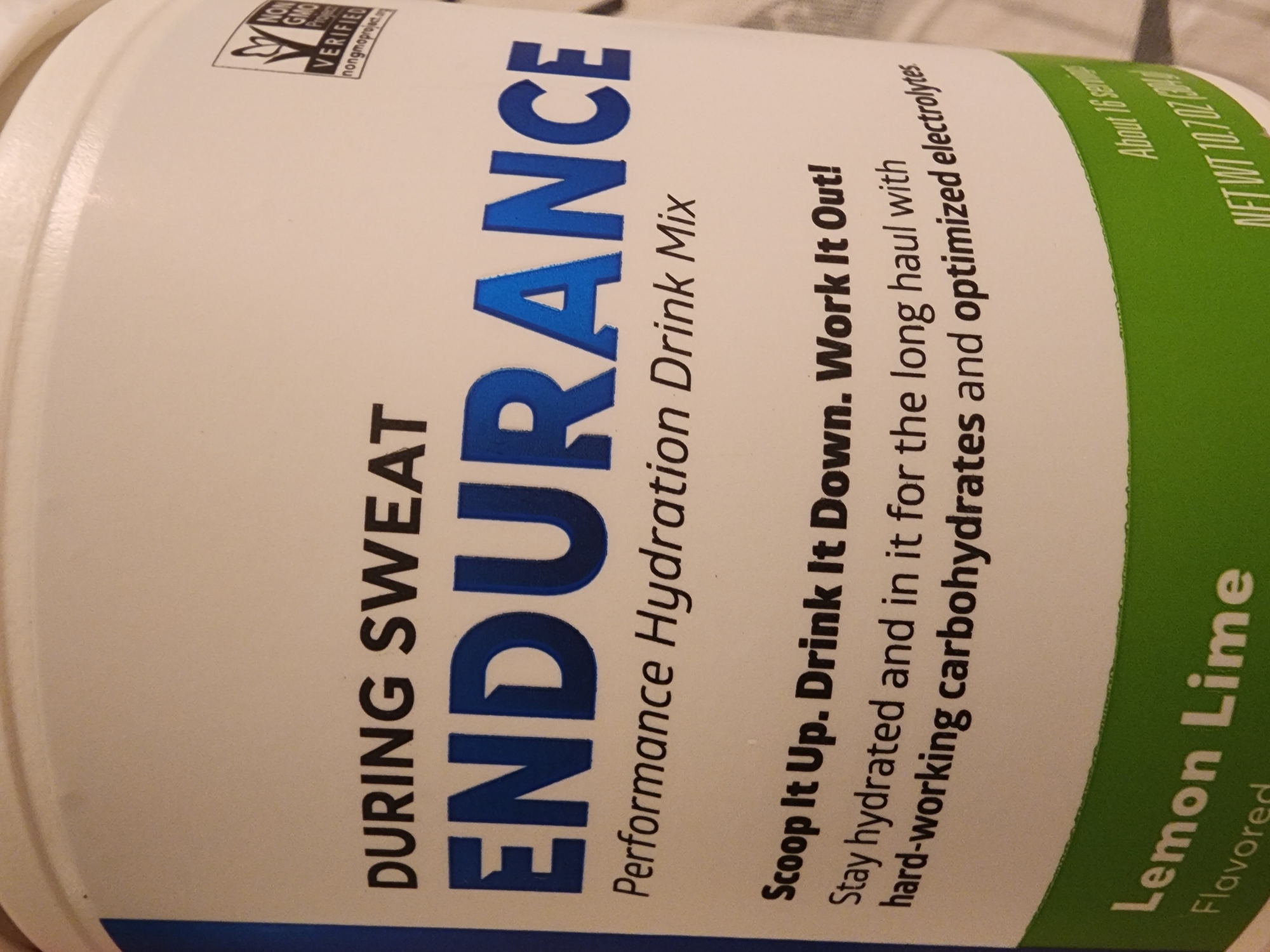 The image is a close-up shot of a white plastic container for "ENDURANCE Performance Hydration Drink Mix" in "Lemon Lime" flavor. The text on the container is predominantly black and blue. The brand name "ENDURANCE" is displayed in large, bold blue lettering. Above it, in black, are the words "DURING SWEAT". Below "ENDURANCE", the words "Performance Hydration Drink Mix" are written in a smaller, cursive-like font. Further down, a tagline reads "Scoop It Up. Drink It Down. Work It Out! Stay hydrated and in it for the long haul with hard-working carbohydrates and optimized electrolytes". A green band runs along the bottom of the container, with the words "Lemon Lime" and "Flavored" visible. A "NON GMO PROJECT VERIFIED" logo is present on the top right of the container. The background appears to be a neutral, possibly fabric surface. There is no discernible information about a specific location in a city.