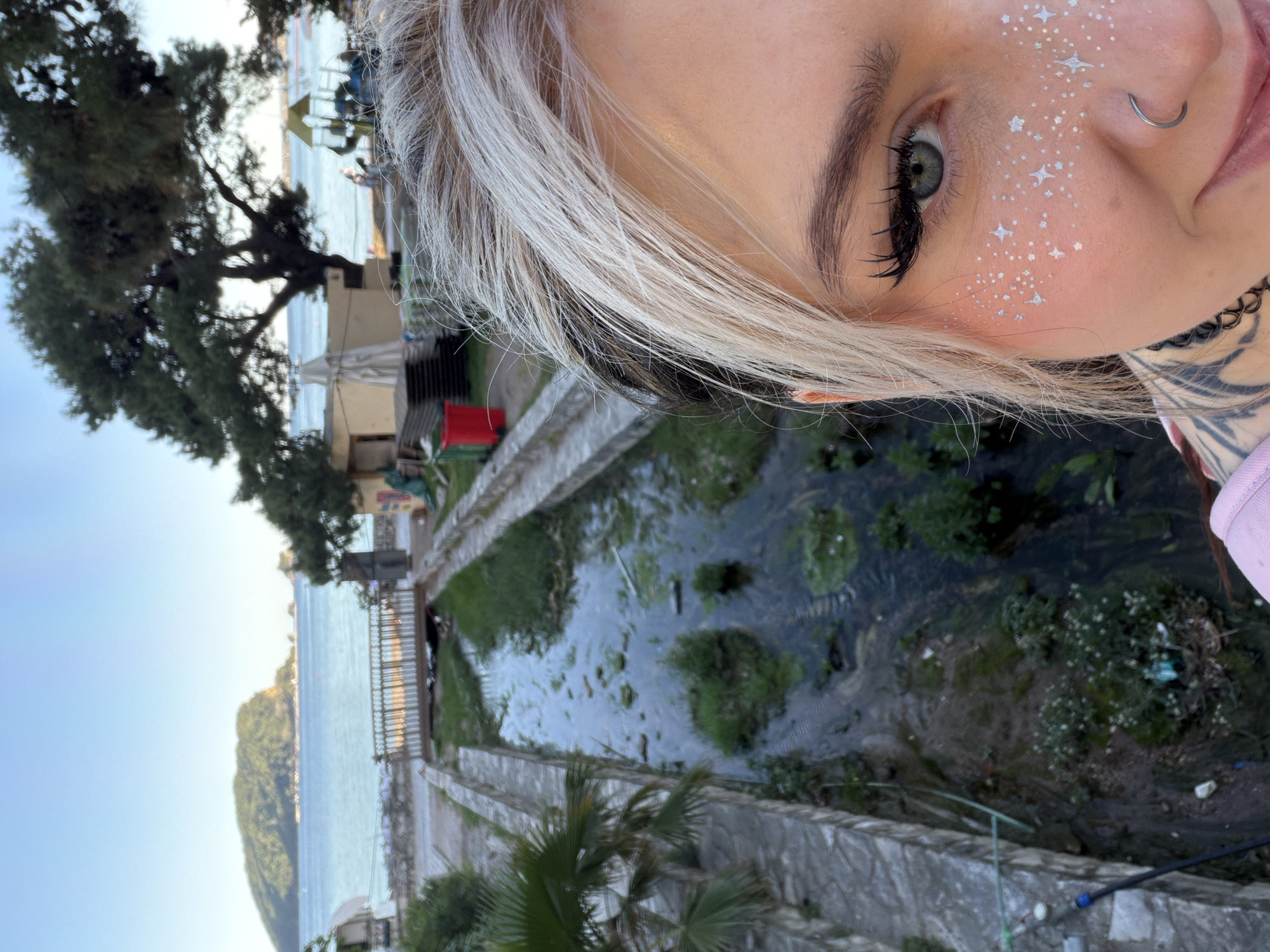 The image captures a selfie taken by a person with platinum blonde hair featuring black roots. The person has eye makeup and white, star-shaped glitter embellishments on the right side of their face. They also have a nose ring and a tattoo visible on their neck. The background reveals an outdoor scene. The person is standing near a retaining wall, with water flowing in what appears to be a canal or drainage system. There are patches of greenery and some debris visible in the water. Beyond the wall and canal, there is a waterfront area with a beach, the sea, and a mountainous island in the distance. The location appears to be in Budva, Montenegro. A building and tree are visible in the background along the water's edge.