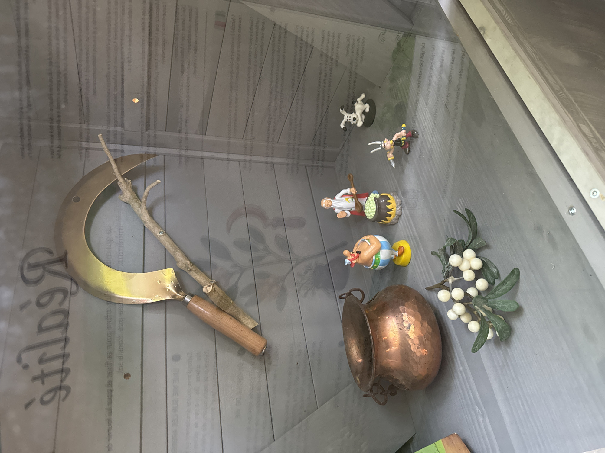 The image shows a display case containing historical agricultural tools and figurines. A golden sickle with a wooden handle is prominently displayed, leaning against a wooden background. To the left of the sickle is a copper pot. In front of the pot, a sprig of white berries on green leaves is arranged. Several small figurines of cartoon characters, likely from the Asterix series, are placed in the display. One figurine of Obelix stands to the right of the copper pot. Further to the right, there are figurines of a man with a white beard stirring a pot, a small white dog, and a character resembling Asterix holding a knife. The background consists of wooden planks with faint text and imagery, suggesting an informational display. The lighting is even, and the overall scene appears to be indoors, possibly in a museum or exhibition. The display is likely located in the Jura Museum of Porrentruy, Switzerland, which features an exhibit on agricultural heritage, including tools and local traditions.