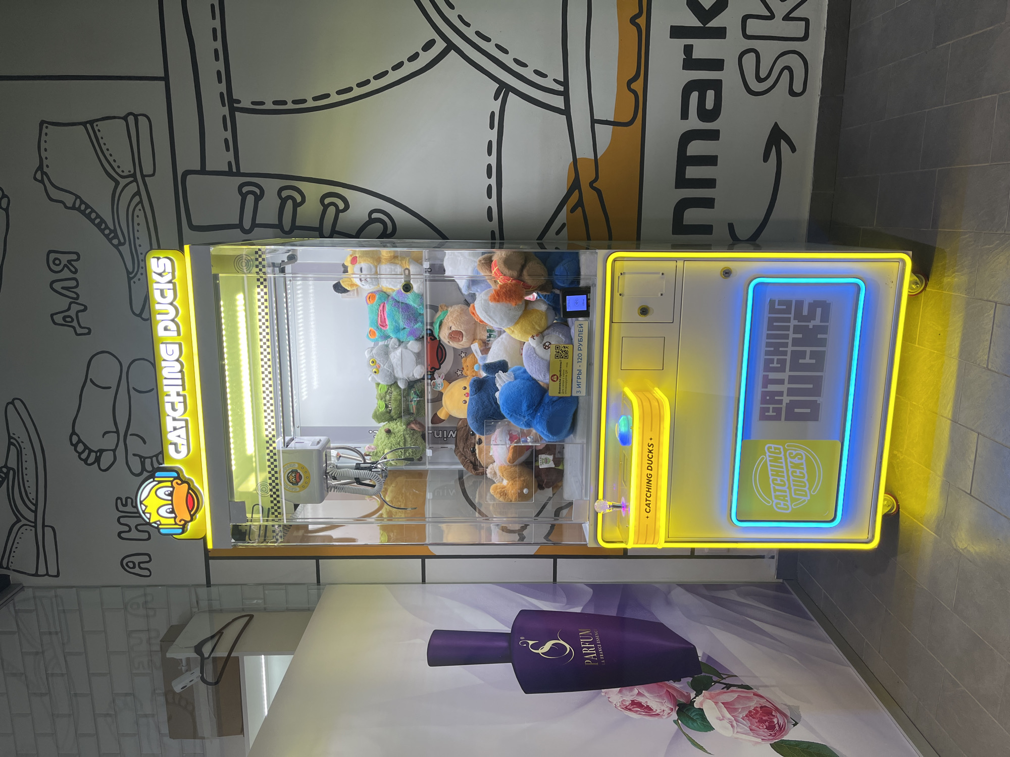 The image shows a "Catching Ducks" claw machine in Izhevsk, Russia. The machine is filled with stuffed animals. Its front panel has a blue-lit screen displaying the game's title, and a yellow sign indicating the game's name and coin insertion slot. Adjacent to the machine is a wall decorated with hand-drawn illustrations of footwear. Below the machine is an advertisement poster featuring a dark purple perfume bottle with floral accents. The flooring appears to be tiled.